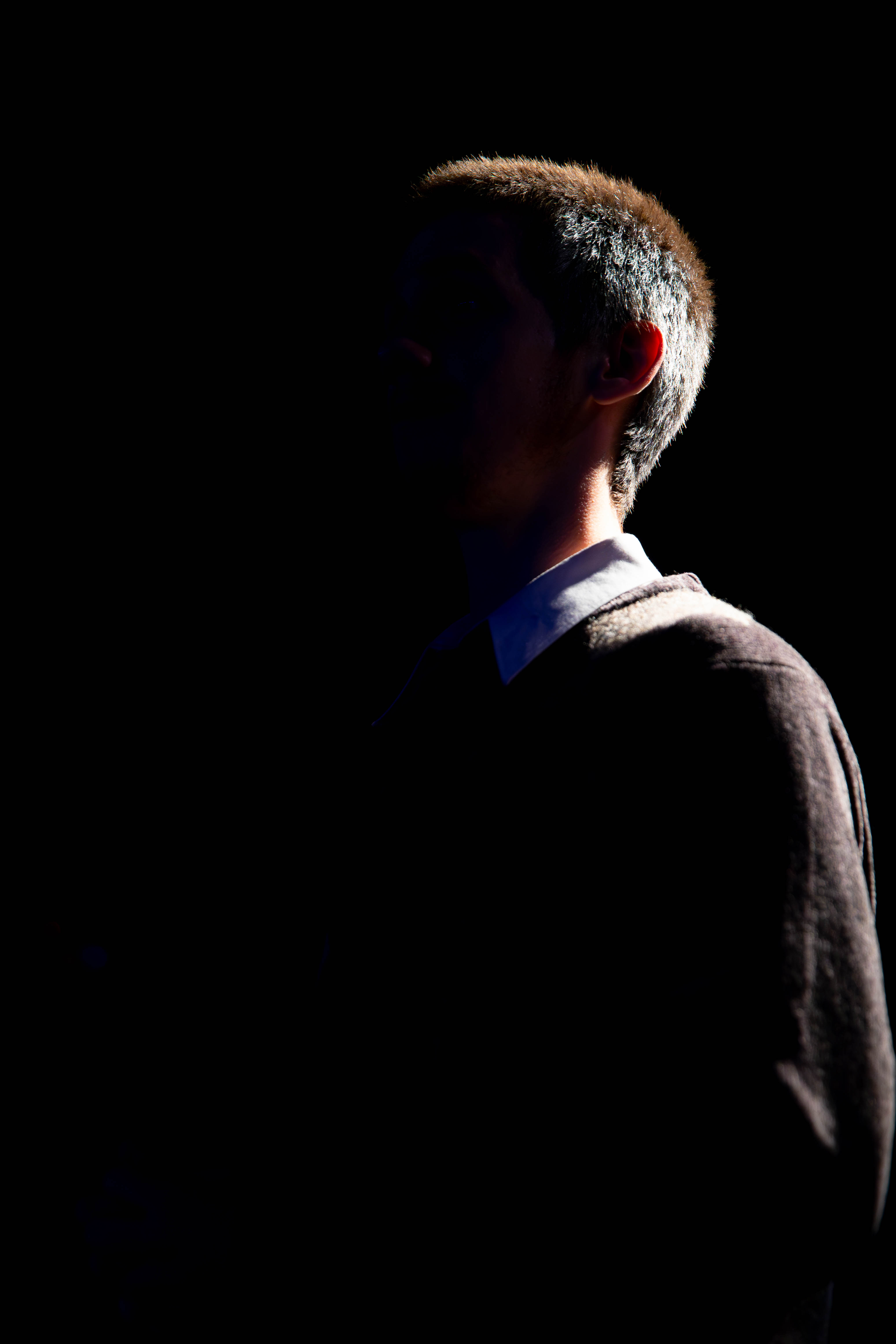 Profile of a person with short hair, wearing a collared shirt and a sweater, illuminated from the side with a dark background.