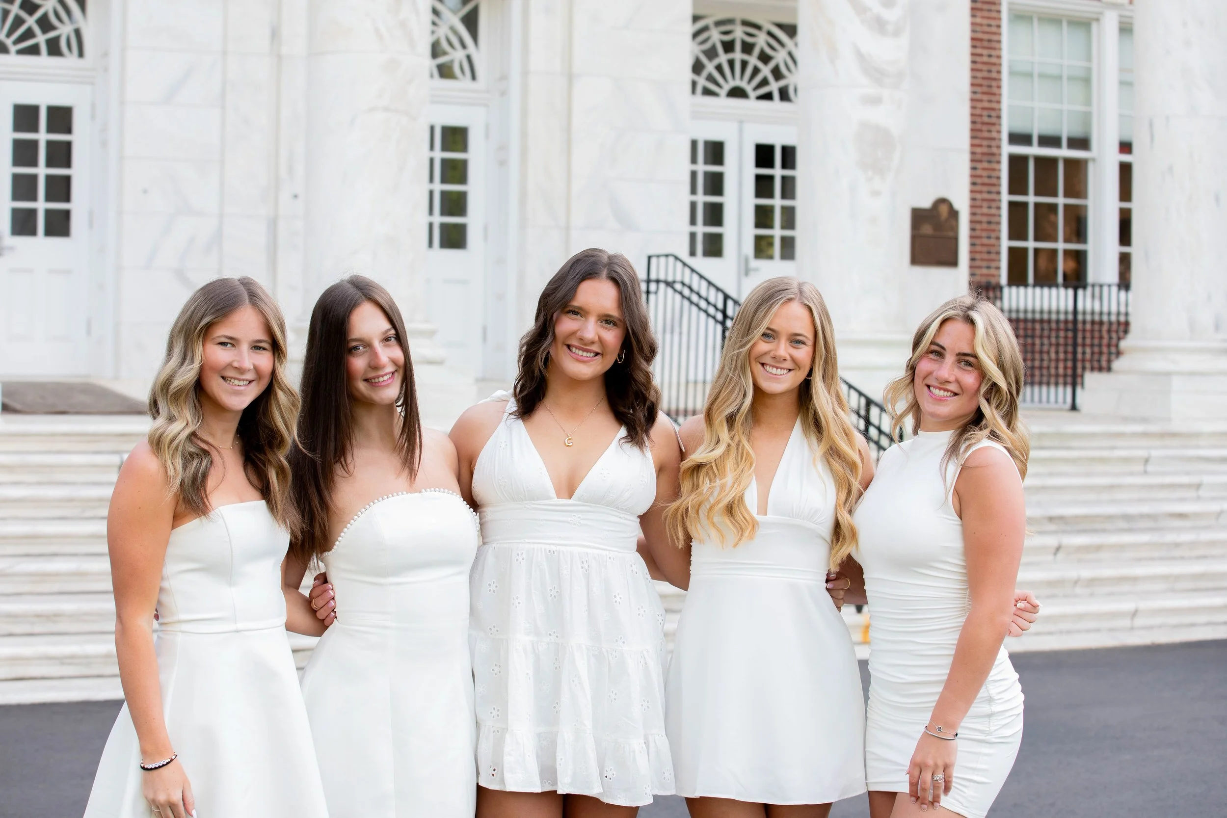 Group of five young women in white dresses standing on the steps of a building with large windows, smiling at the camera.