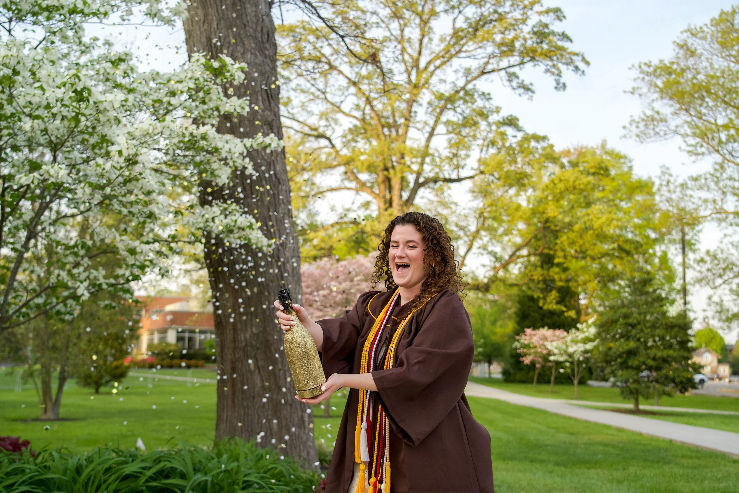 A woman in a graduation gown and cap smiling and celebrating outdoors while popping open a bottle of champagne, with trees and a park in the background.
