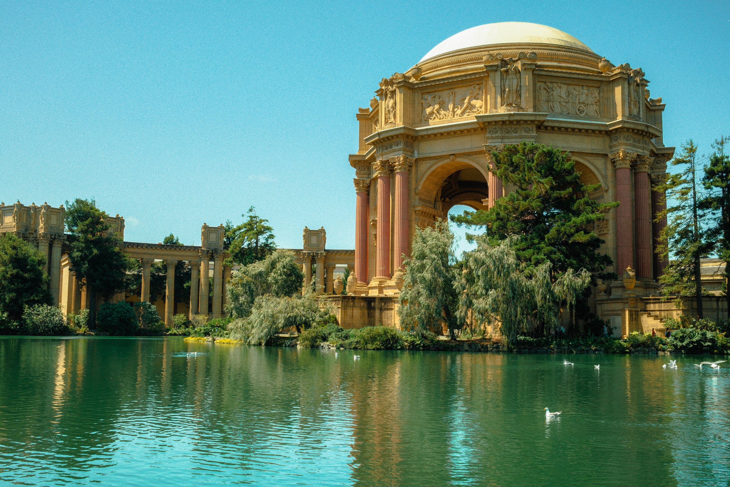A historic seaside building with elaborate classical architecture, featuring columns, sculptures, and a domed roof, surrounded by trees and reflected in a body of water.