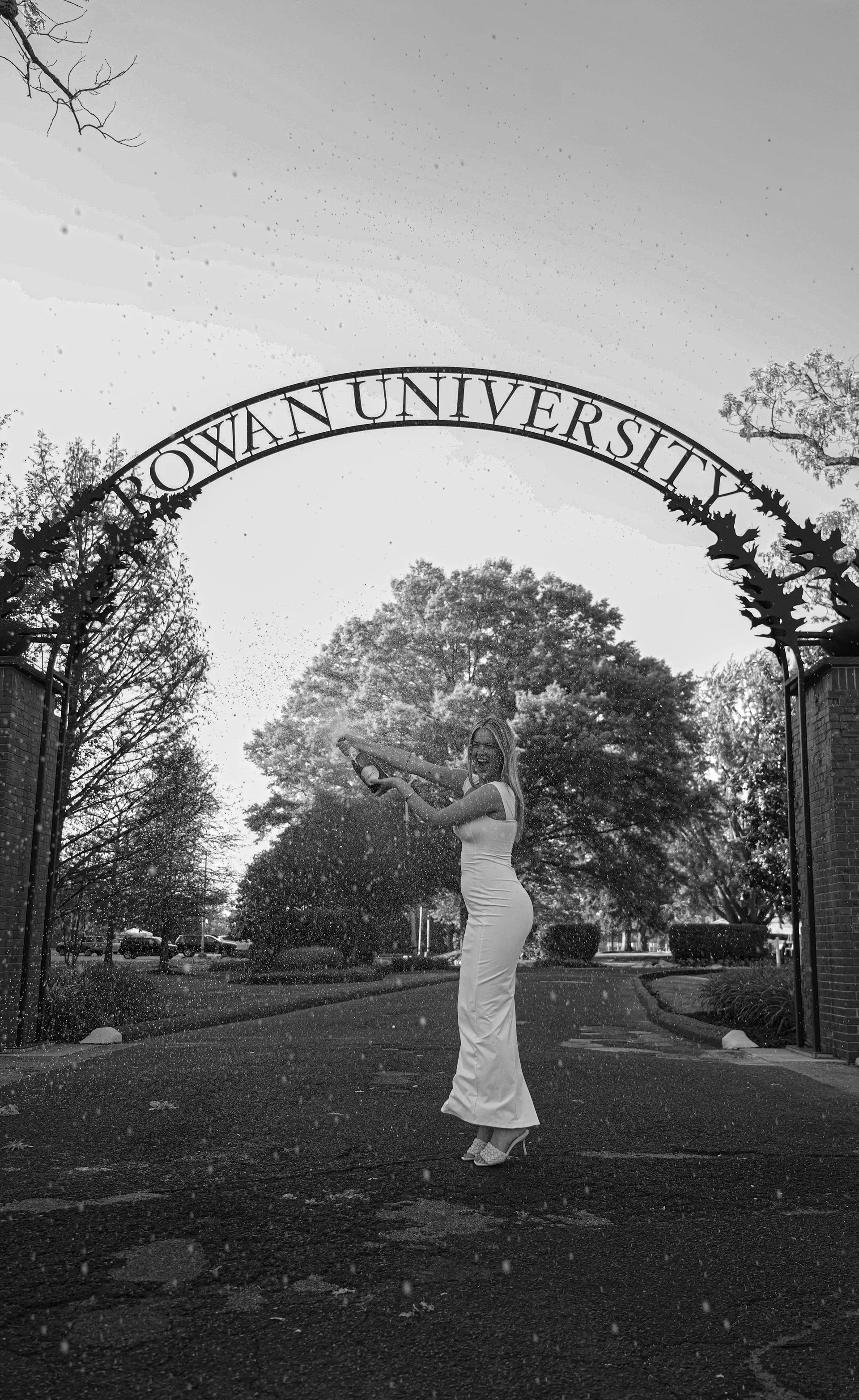 A woman in a white dress and high heels is smiling and spraying champagne under the archway that reads "OWA UNIVERSTY" in a park area with trees and cars in the background.