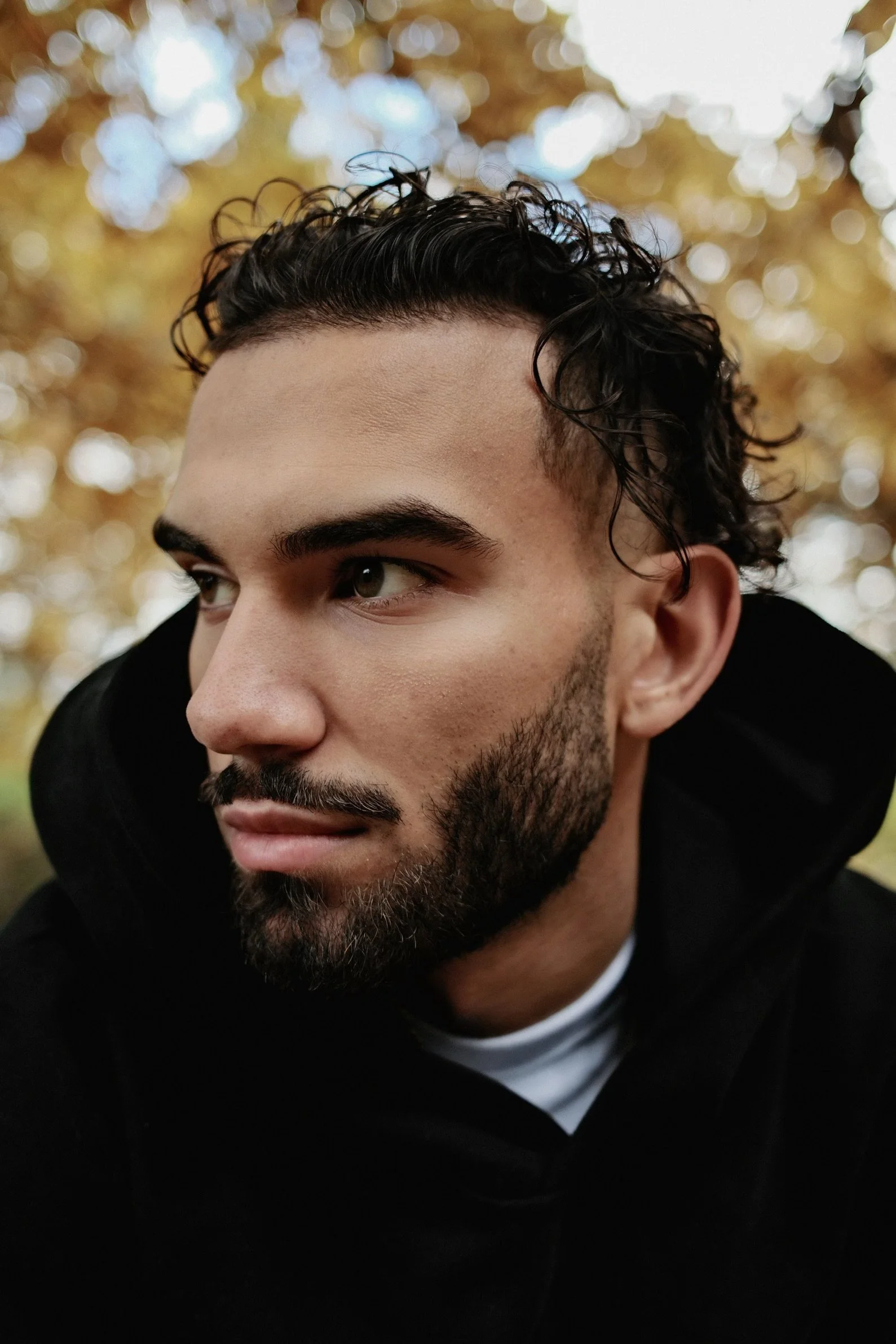 A close-up of a man with dark curly hair and a beard, wearing a black hoodie, looking to the right, with a background of golden and blue blurred lights.