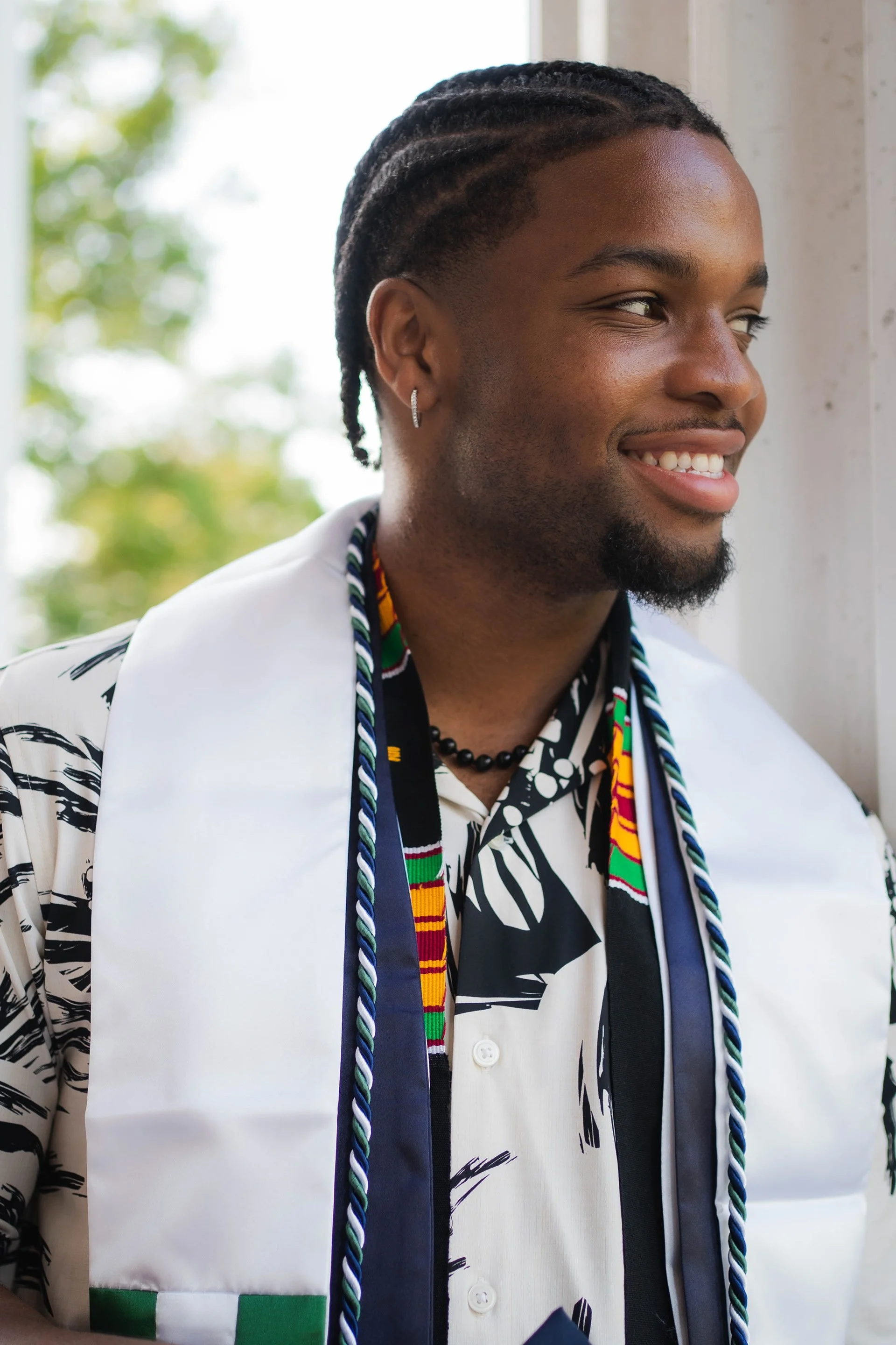 A young man with dark braided hair, earrings, and a beard, smiling and looking to the right, wearing a colorful scarf, patterned shirt, and a white jacket, standing near a window with blurred green foliage in the background.