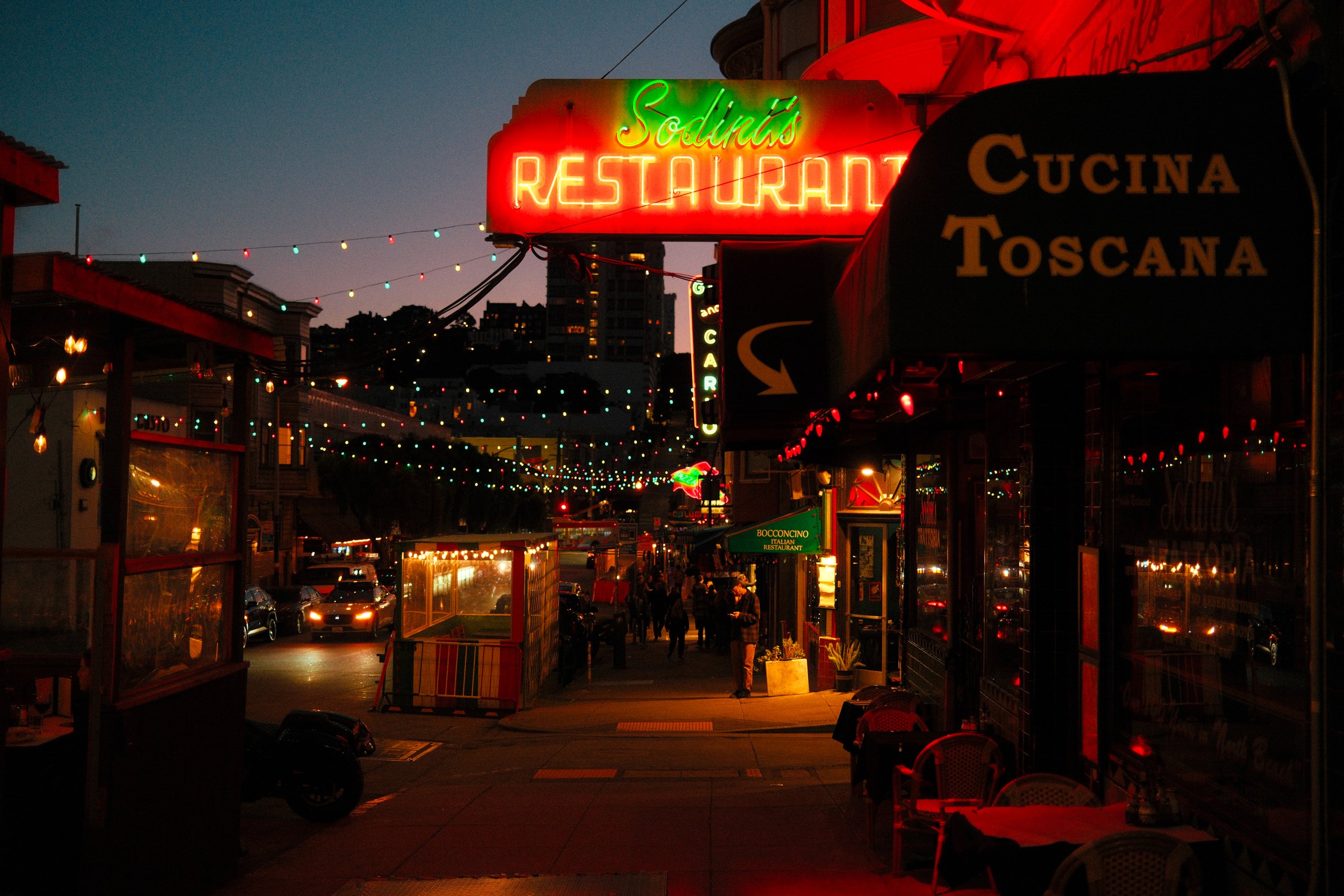 Night scene of a lively street with illuminated restaurant signs, including a neon sign that reads 'Soluma's Restaurant' and a smaller sign for 'Cucina Toscana'. String lights decorate the street area, and there are people walking along the sidewalk.