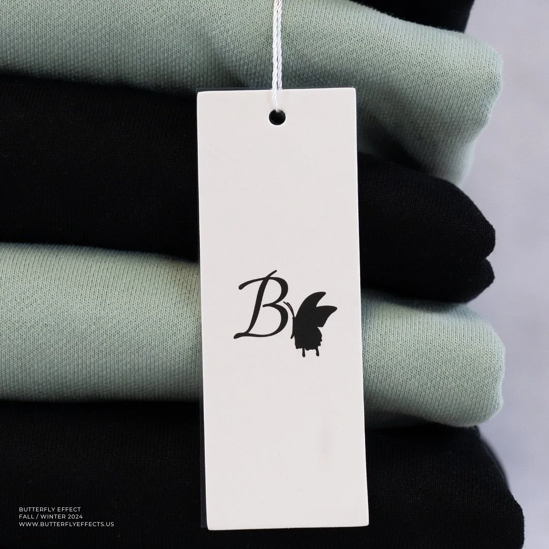 Stacked folded clothing in light green, black, light green, and black with a white price tag displaying the word "B" and a butterfly logo.