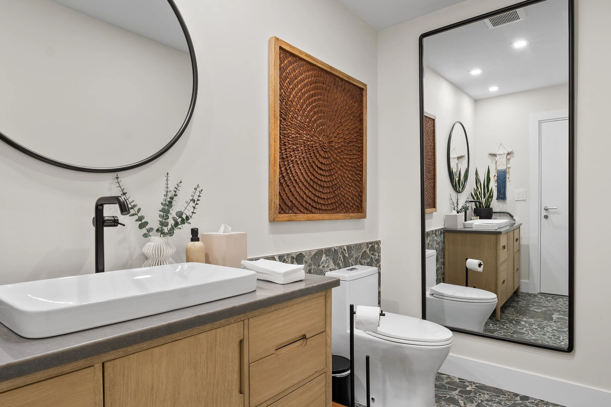 Bathroom with white sink, wooden vanity, mirror, bathroom artwork, plants, toilet, toilet paper, and a shower with a glass door, white walls, and ceiling lights.