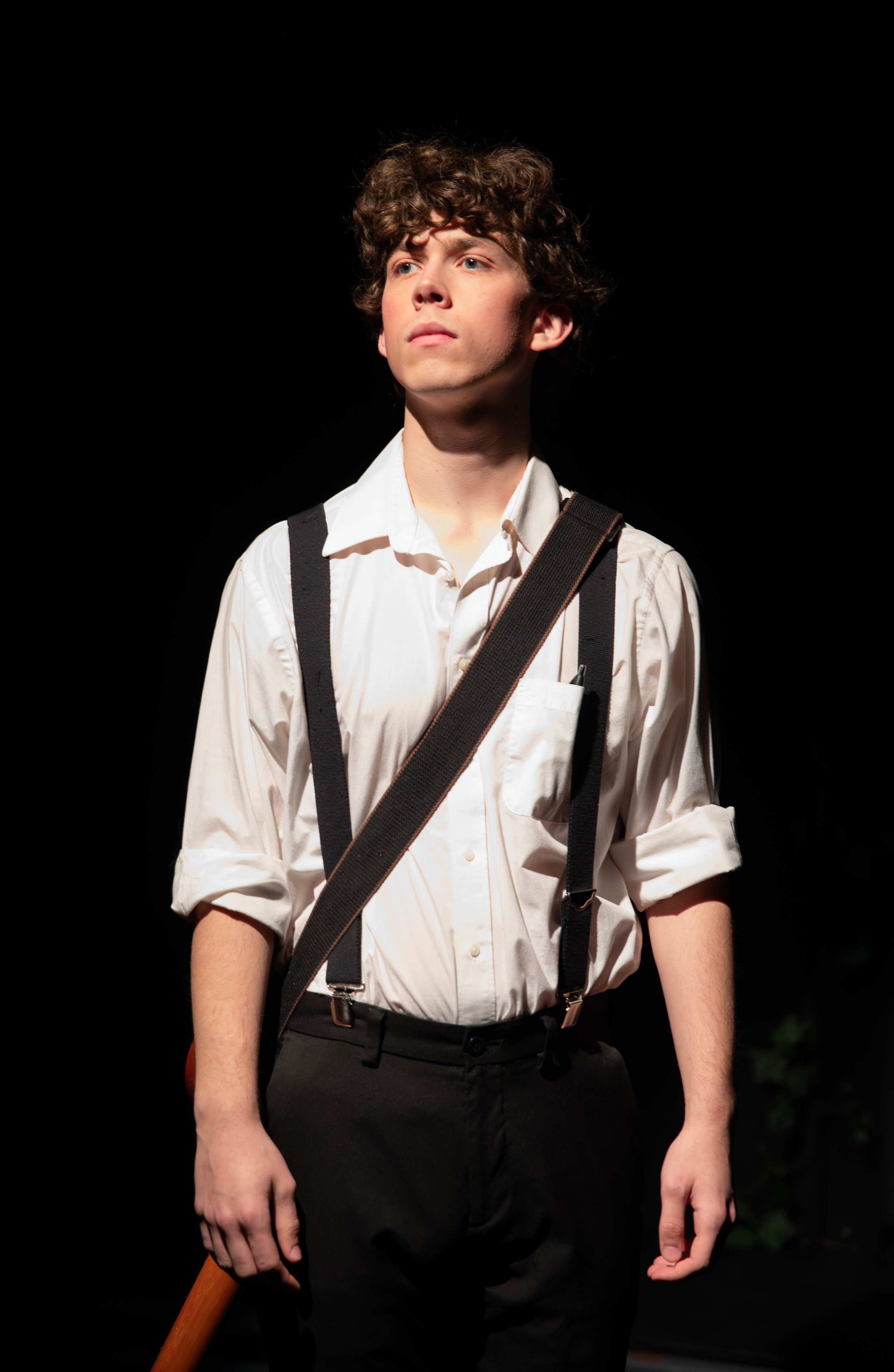 A young man with curly brown hair wearing a white dress shirt, suspenders, and black pants, standing against a dark background. He has a serious expression, looking slightly upward, with a brown baseball bat resting against his side.