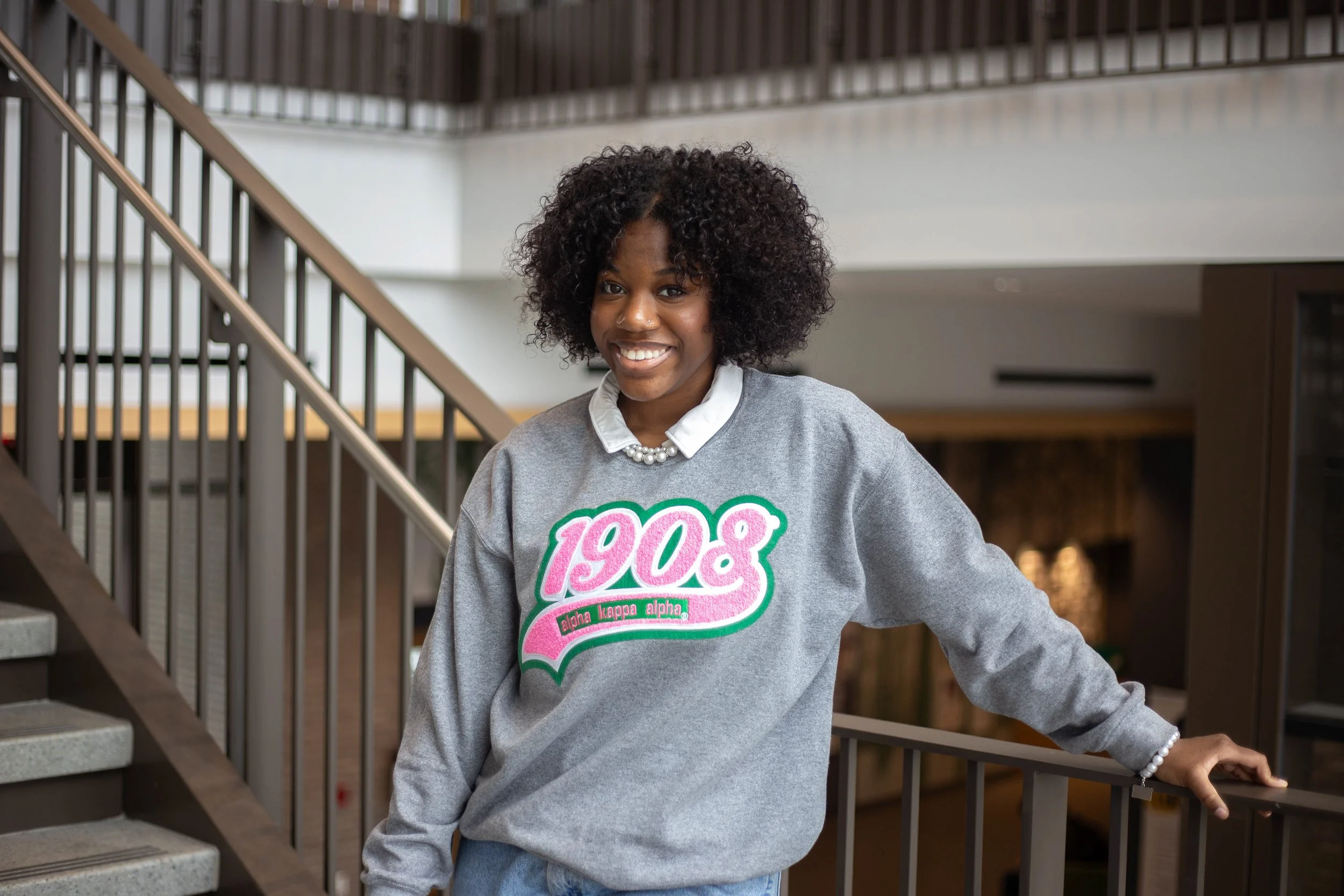 A young woman with curly hair smiling while standing on a staircase inside a building. She is wearing a gray sweatshirt with pink and green text and a white pearl necklace.