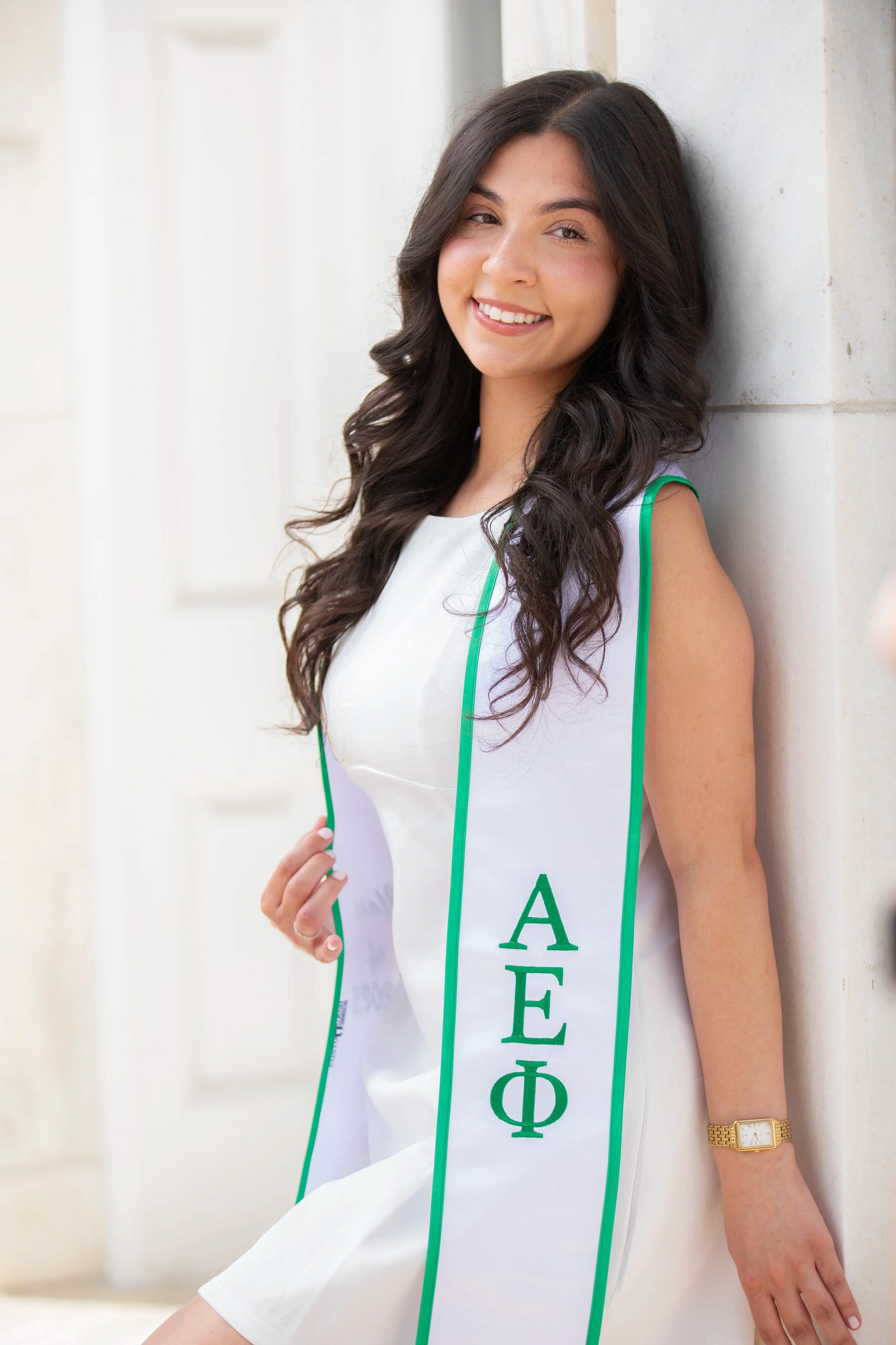 Young woman with long dark wavy hair smiling, wearing a white dress and a gold watch, leaning against a light-colored wall, wearing a sash with Greek letters.