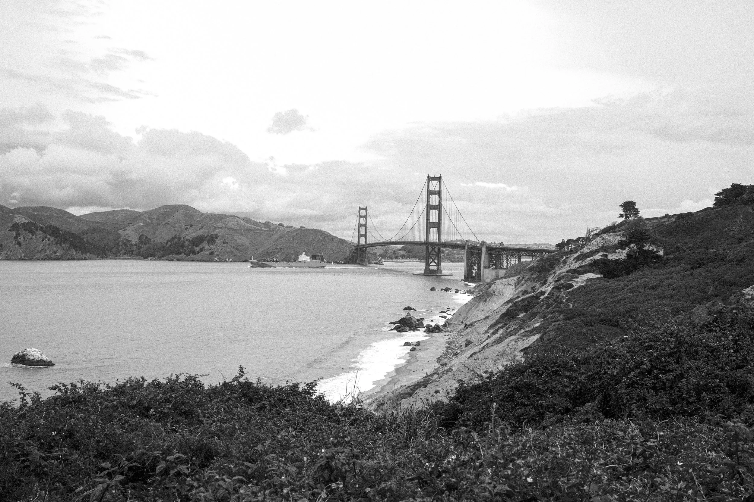 Black and white photo of the Golden Gate Bridge in San Francisco surrounded by hills and water, with some boats visible.