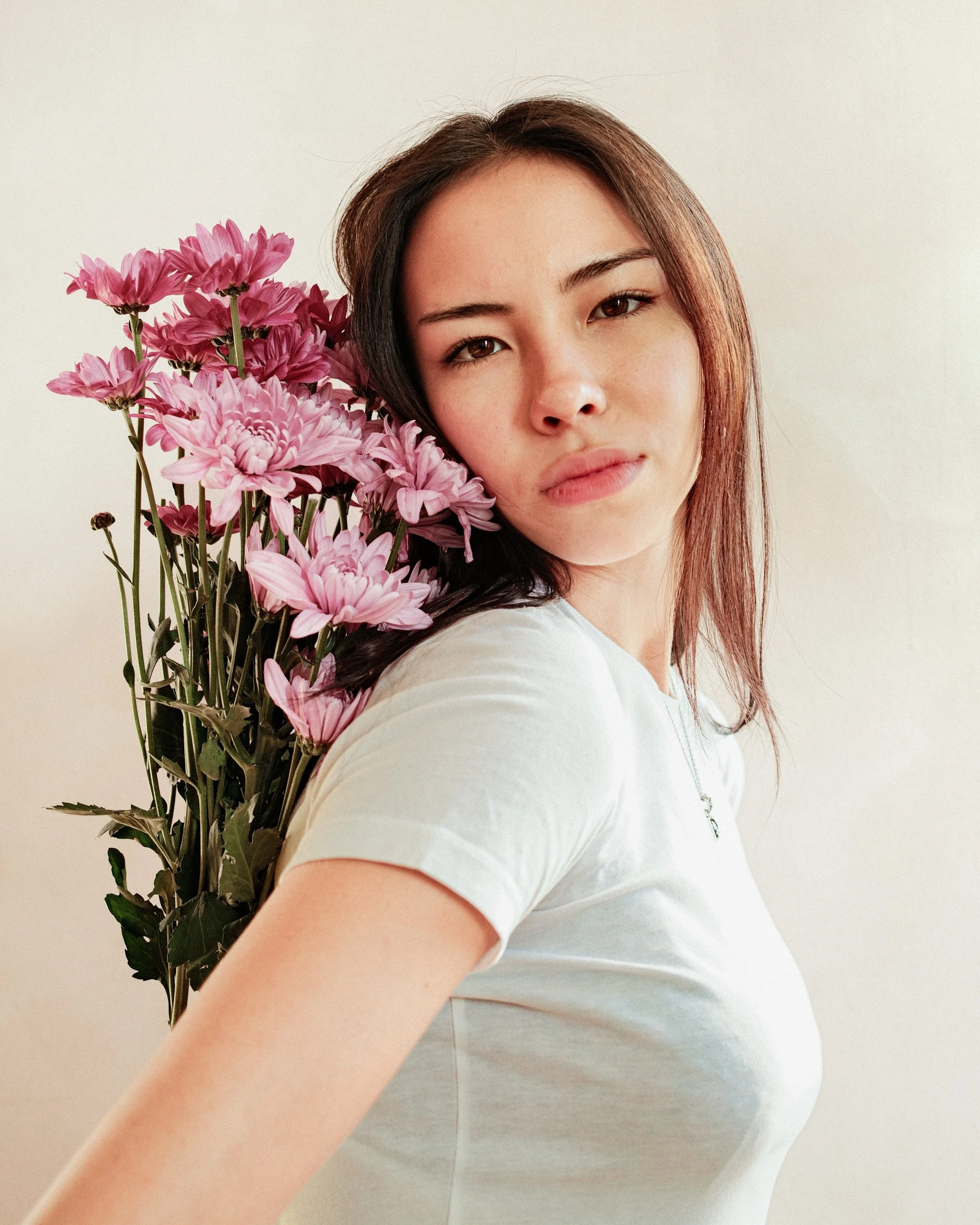 A young woman with brown hair holding pink flowers, wearing a white T-shirt and a necklace with a cross pendant, standing against a plain light background.