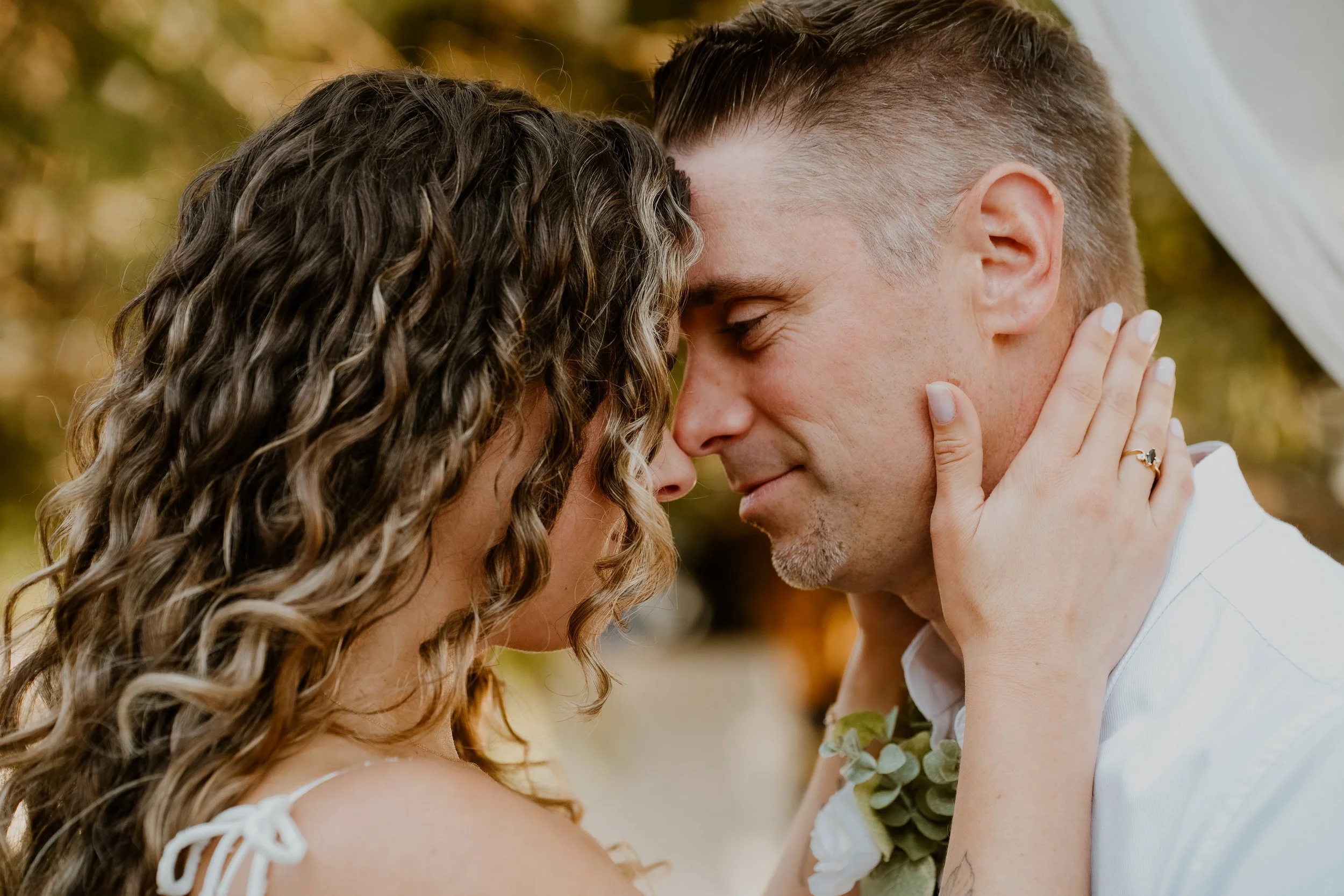A couple sharing a close, intimate moment with foreheads and noses touching, eyes closed, outdoors with autumn foliage in the background.