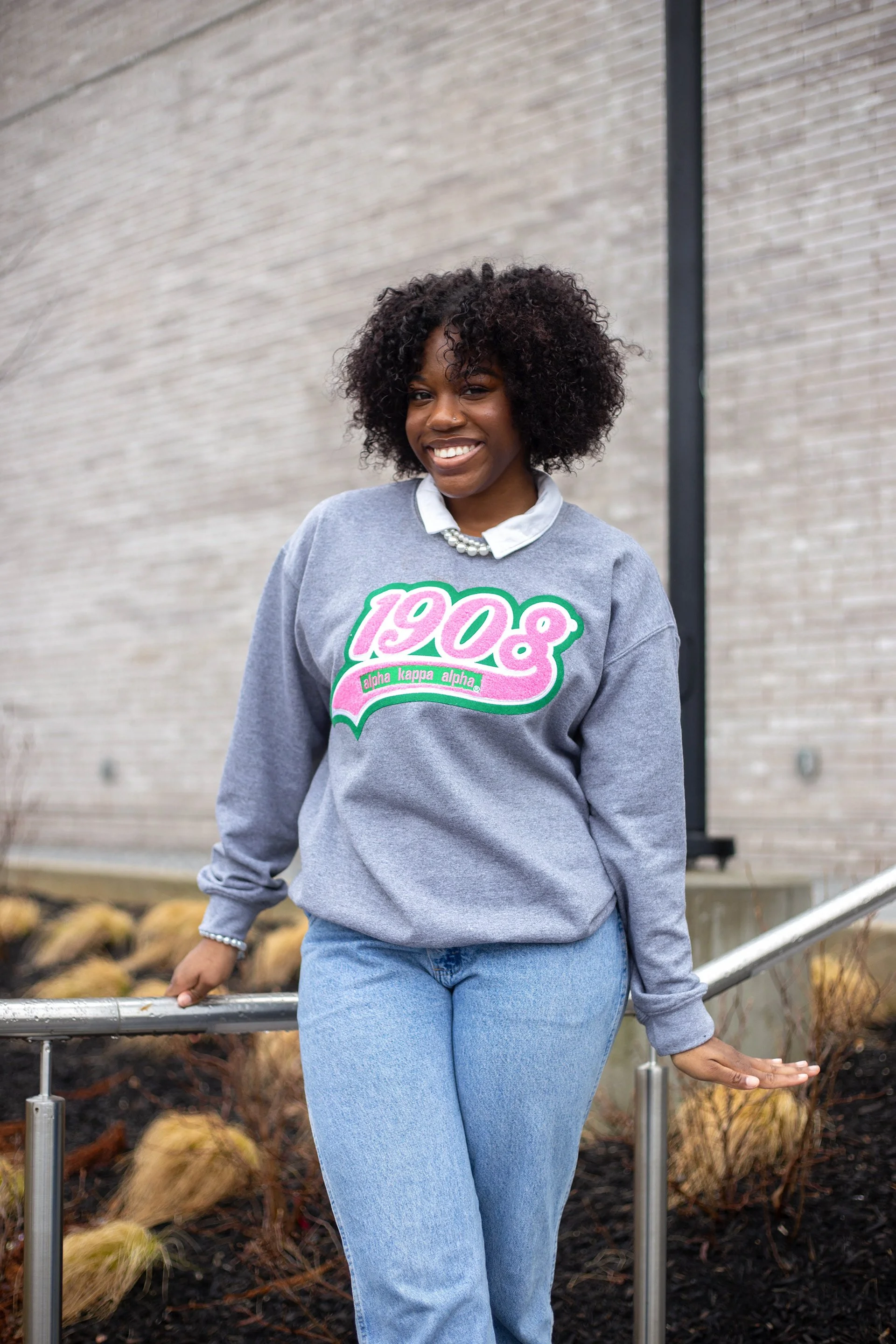 A woman with dark curly hair, wearing a gray sweatshirt with pink and green lettering, standing outdoors near a brick wall and metal railing, smiling at the camera.