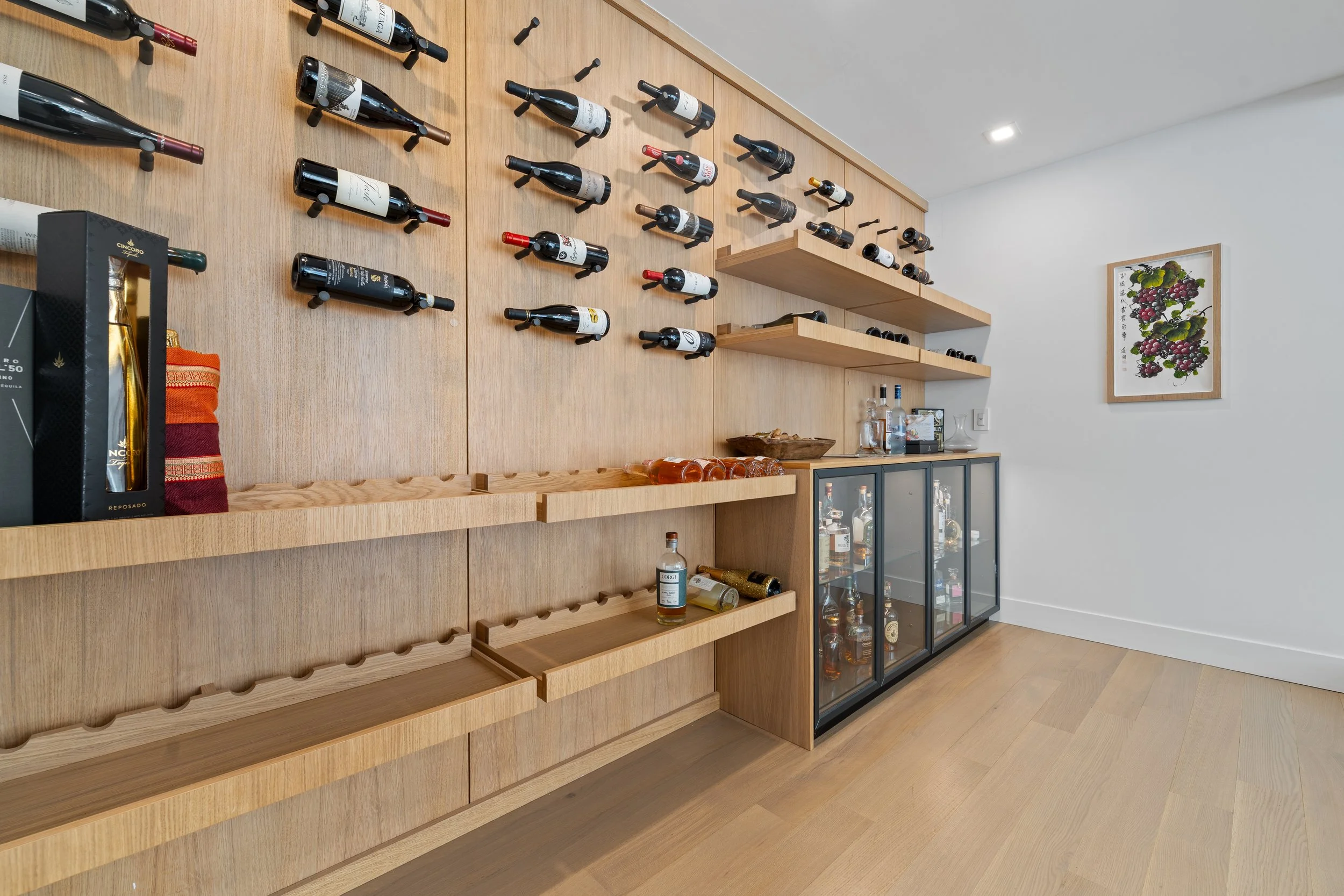 Interior of a wine bar or restaurant with a wooden wall displaying multiple wine bottles in horizontal slots. A glass cabinet with bottles and glasses is below the wall, and a framed artwork featuring grapevines hangs on a white wall to the right.