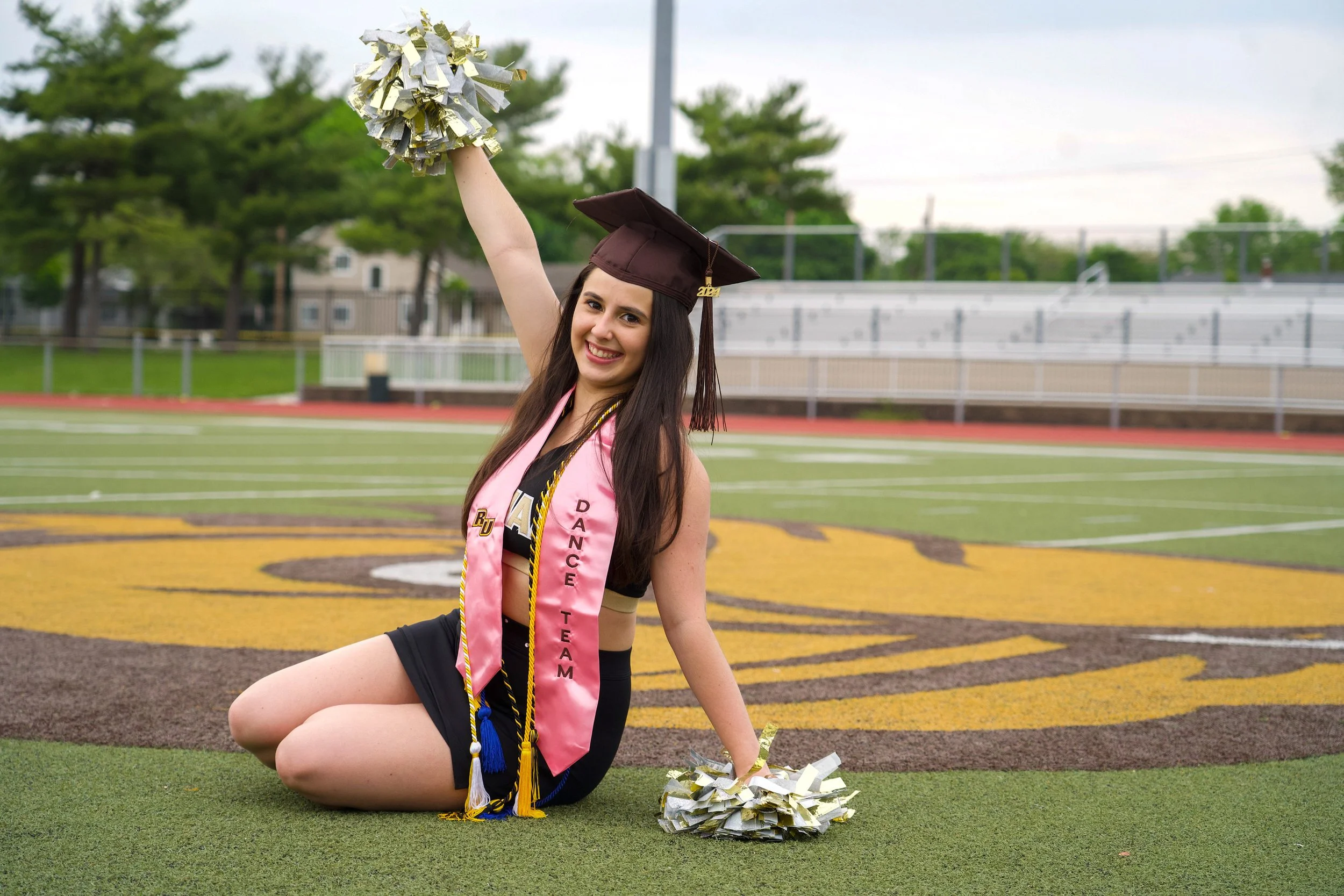 A young woman in a graduation cap and gown sitting on a football field, smiling, holding a pom-pom in her hand with a graduation stole and cords around her neck.