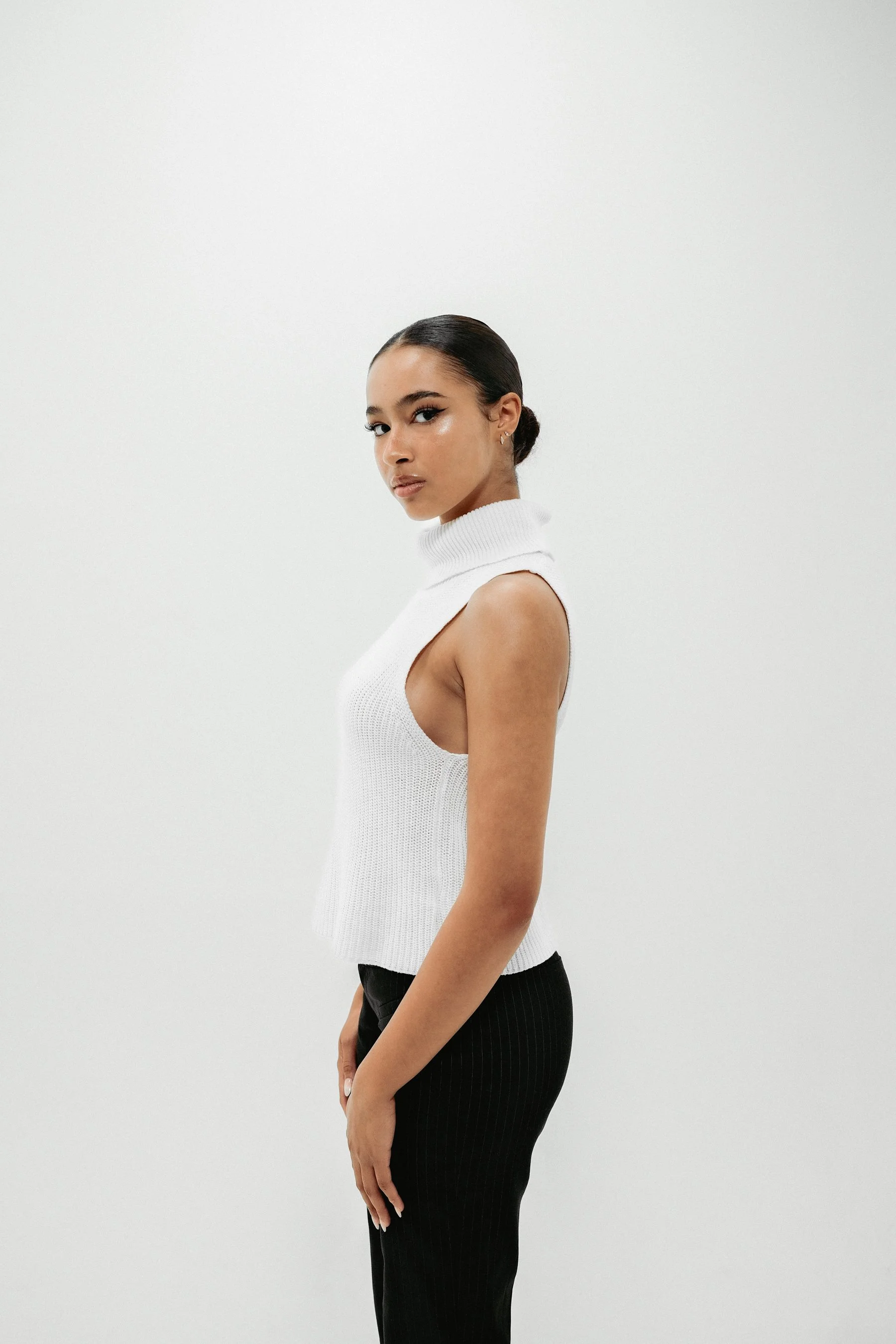 A woman with dark hair styled in a bun, wearing a sleeveless white turtleneck sweater and black pants, posing against a plain white wall.