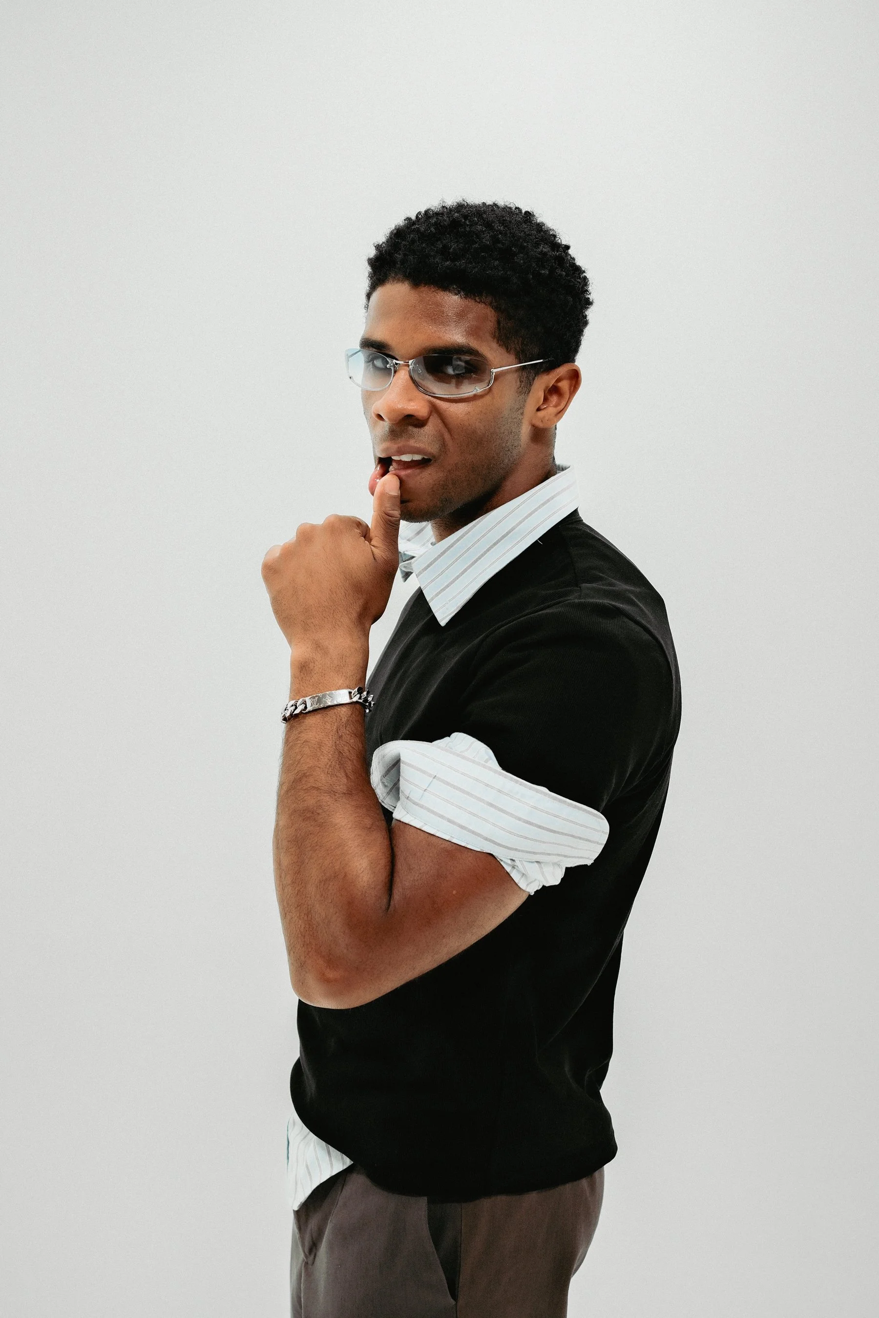 A man with dark curly hair wearing sunglasses, a black t-shirt with rolled-up sleeves over a white striped shirt, a silver bracelet, and a watch, posing against a plain white background, with his left hand on his chin.