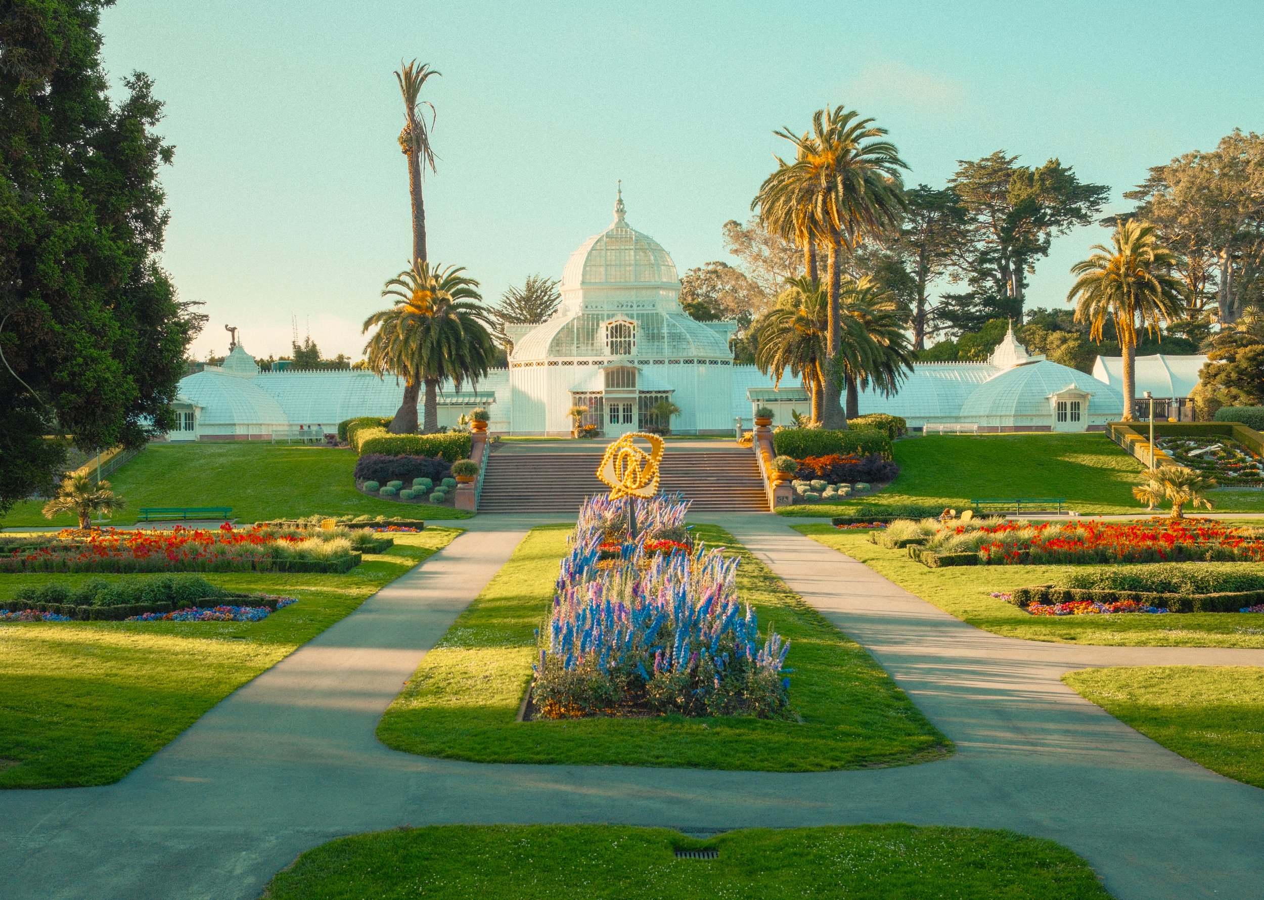 A Victorian-style greenhouse with white walls and domed glass roofs, surrounded by manicured gardens and tall palm trees, under a clear sky.