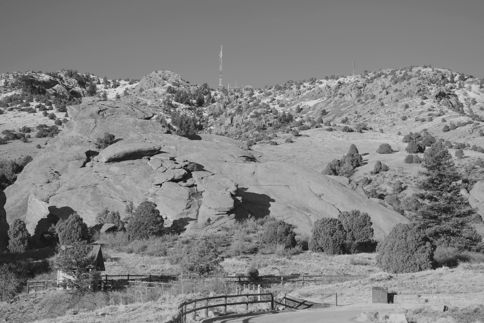 Black and white photo of a mountainous landscape with large rocks and scattered bushes. A fence runs along the foreground, and there are hills with sparse trees and communication towers in the background under a clear sky.