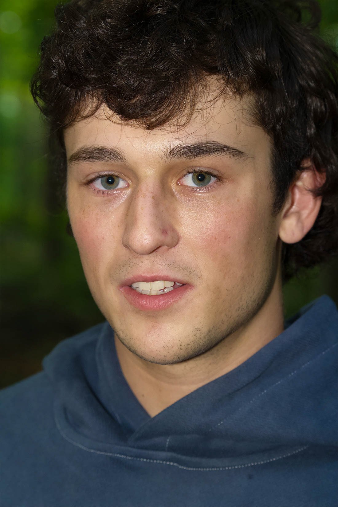 Close-up of a young man's face with curly dark hair, blue eyes, light skin with freckles, wearing a dark blue hoodie, outdoors with blurry green background.