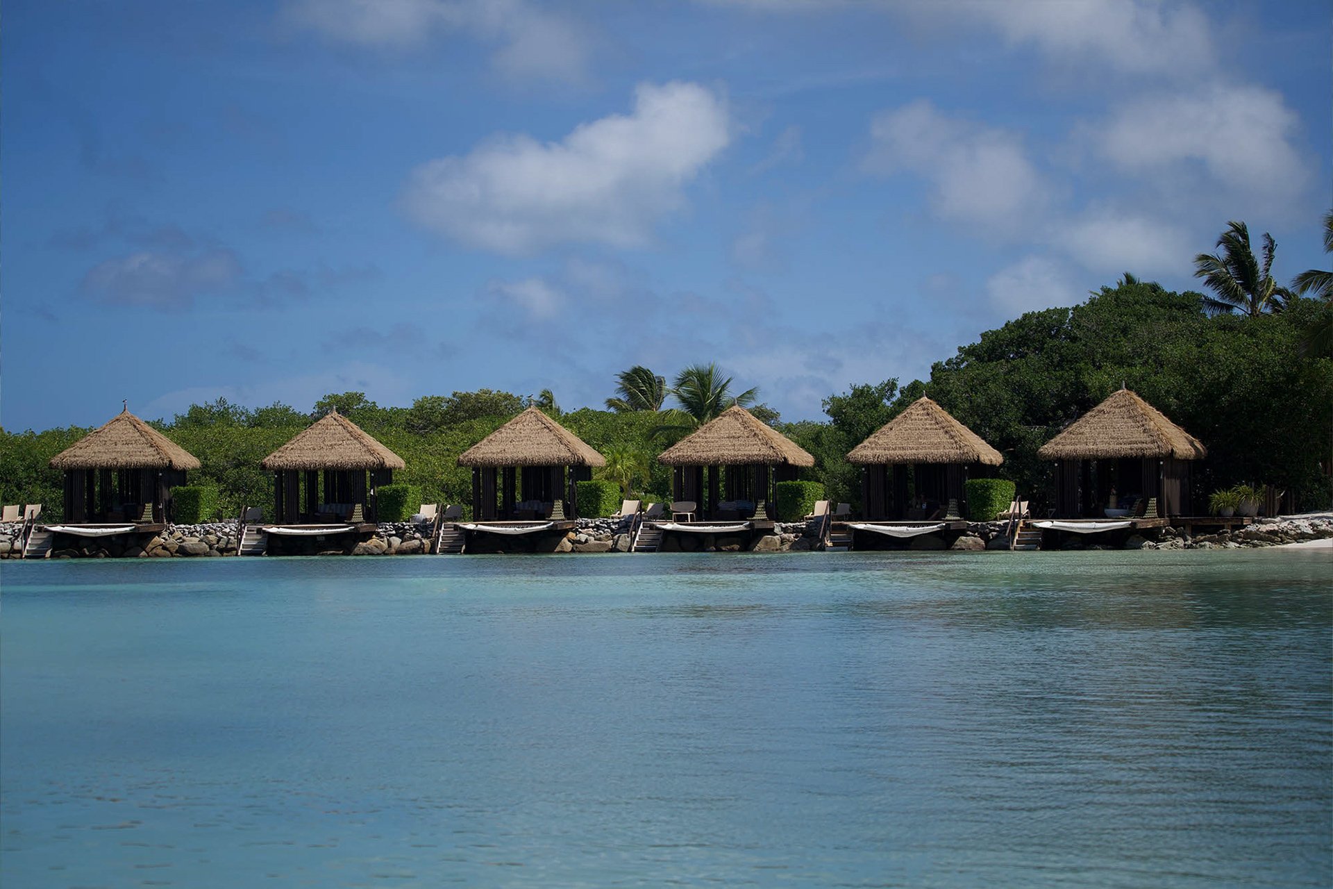 A row of thatched-roof bungalows with lounge chairs on a rocky shoreline, surrounded by tropical trees, under a partly cloudy sky.
