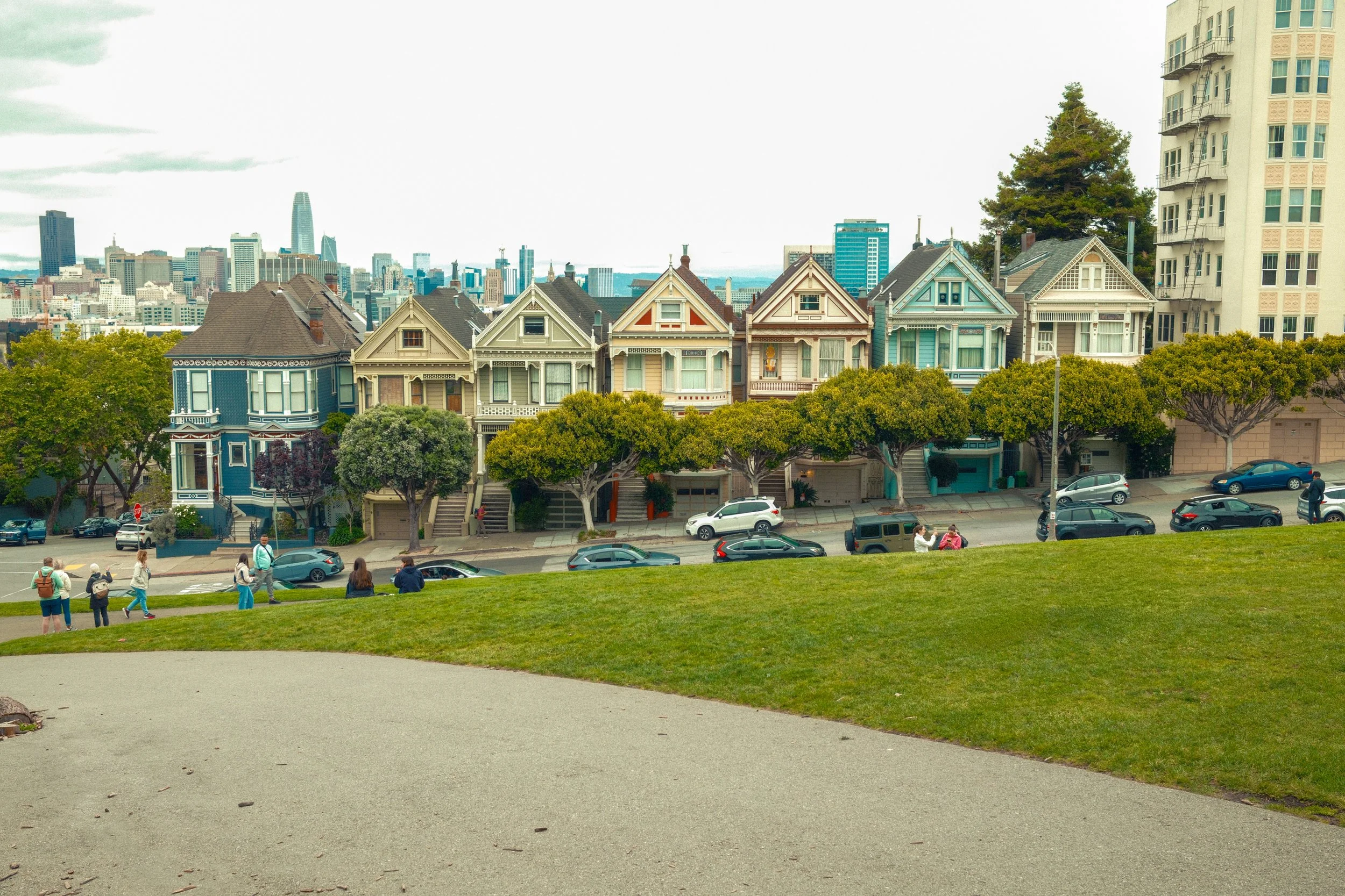 View of Alamo Square with row of Victorian houses, park, and the San Francisco skyline in the background, under cloudy sky.