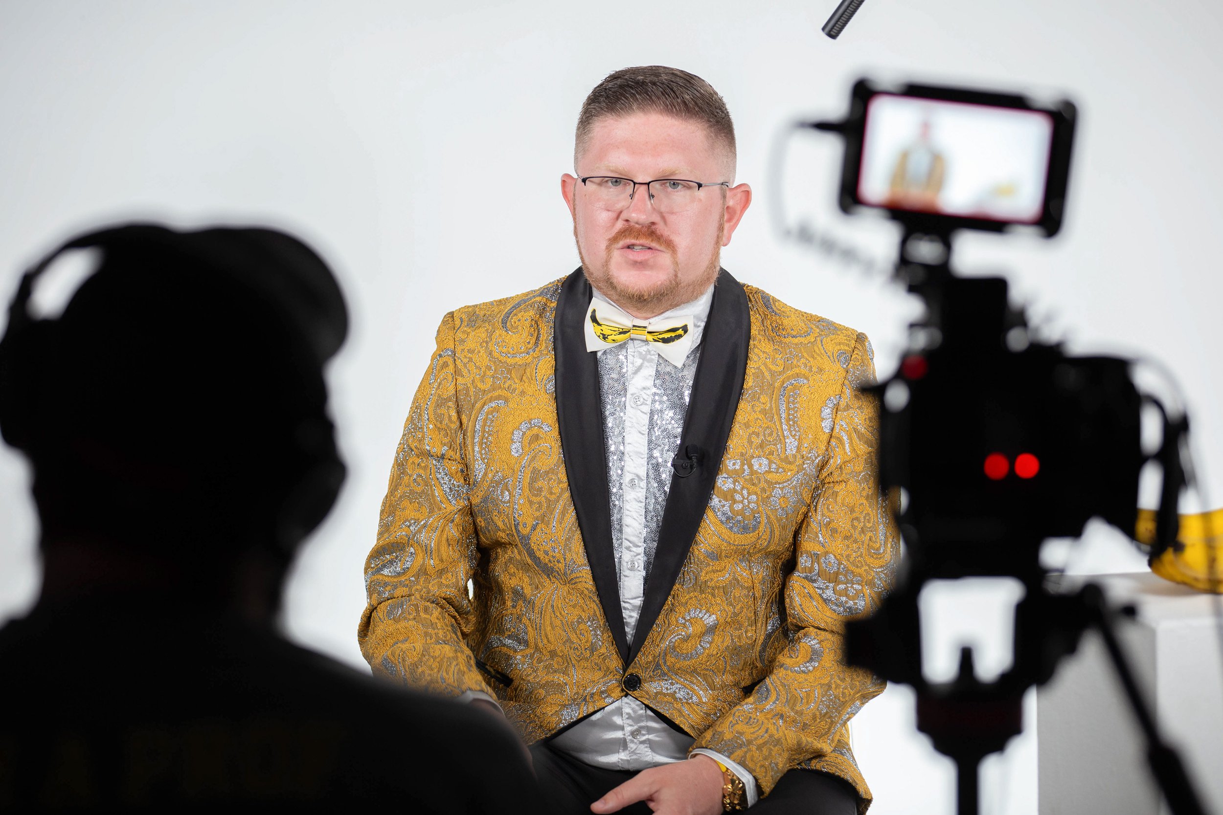 A man with glasses and a beard wearing a gold patterned tuxedo jacket with black lapel and a white shirt, sitting against a plain white background, being recorded by multiple cameras.