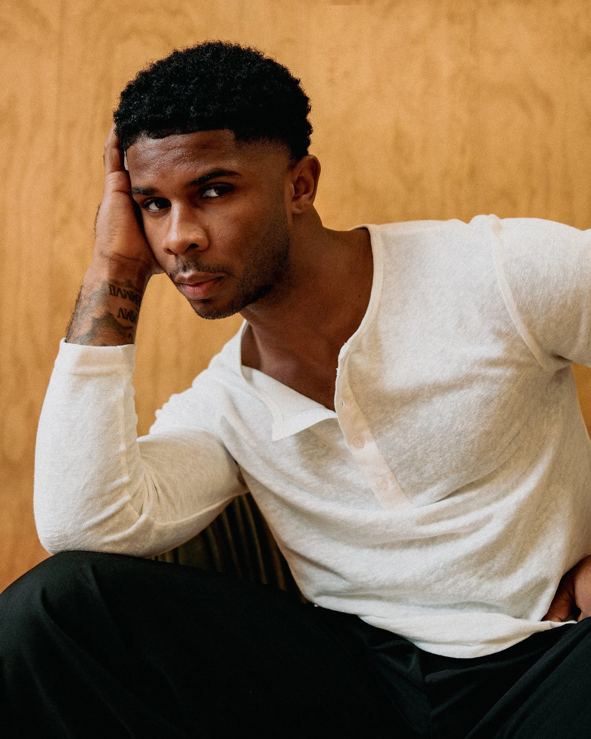 A young man sitting with his head resting on his hand, looking into the camera. He has short, curly black hair, a slight beard, and is wearing a white shirt and black pants. The background is a plain wooden wall.