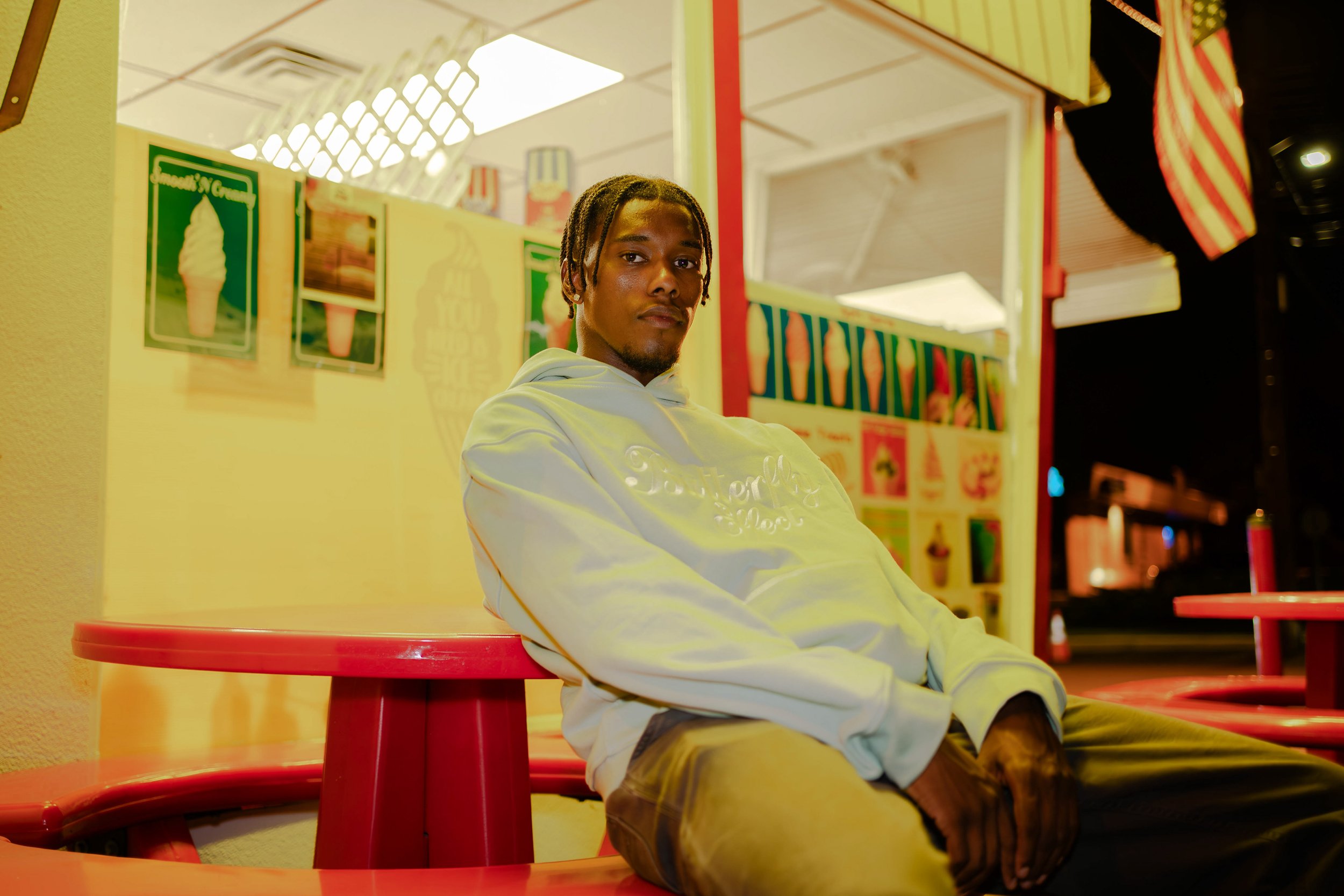 A young man with braided hair sitting at a red table outside a colorful ice cream shop at night.