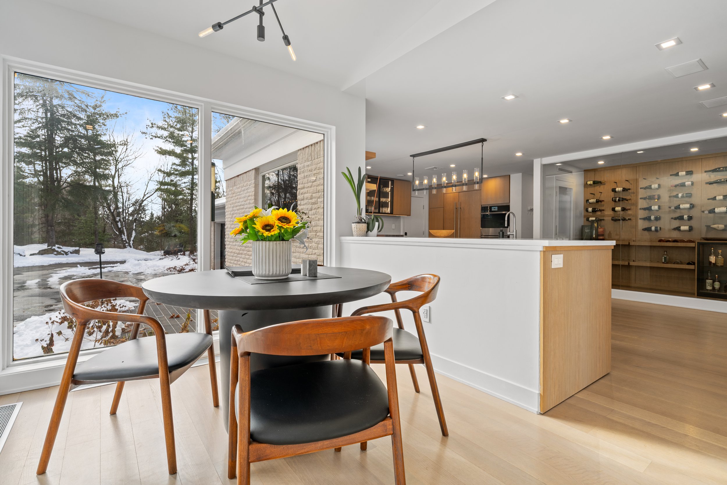 Interior of a modern kitchen and dining area with a white ceiling and walls, wood cabinets, and a wine display on the wall. There is a round dining table with a vase of sunflowers, surrounded by mid-century modern chairs. Large windows show a snowy l