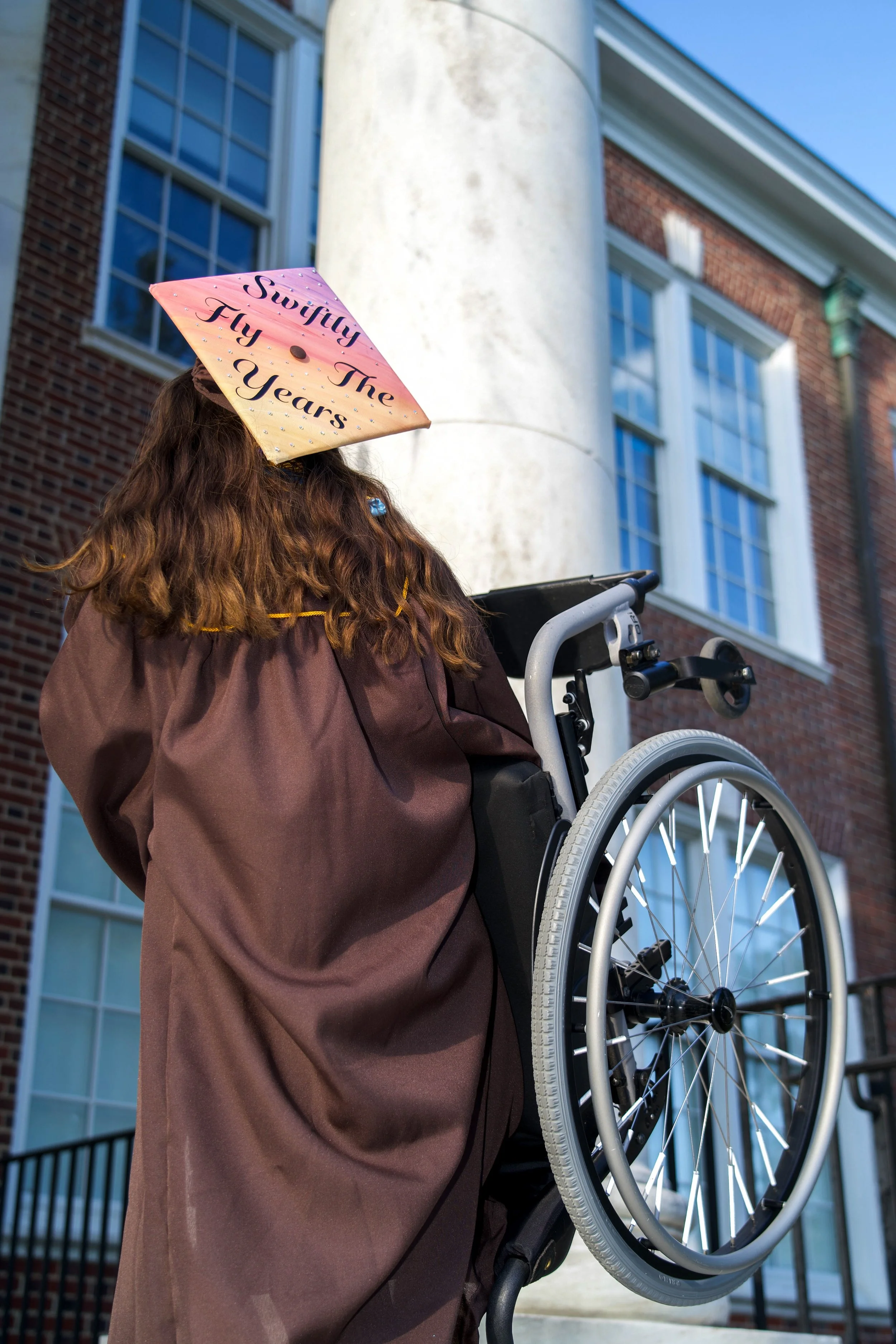 A woman in a brown gown and a pink diploma cap with 'Swifty The Fly 10 Years' written on it, standing outside near a brick building with large windows, holding a wheelchair with large wheels.