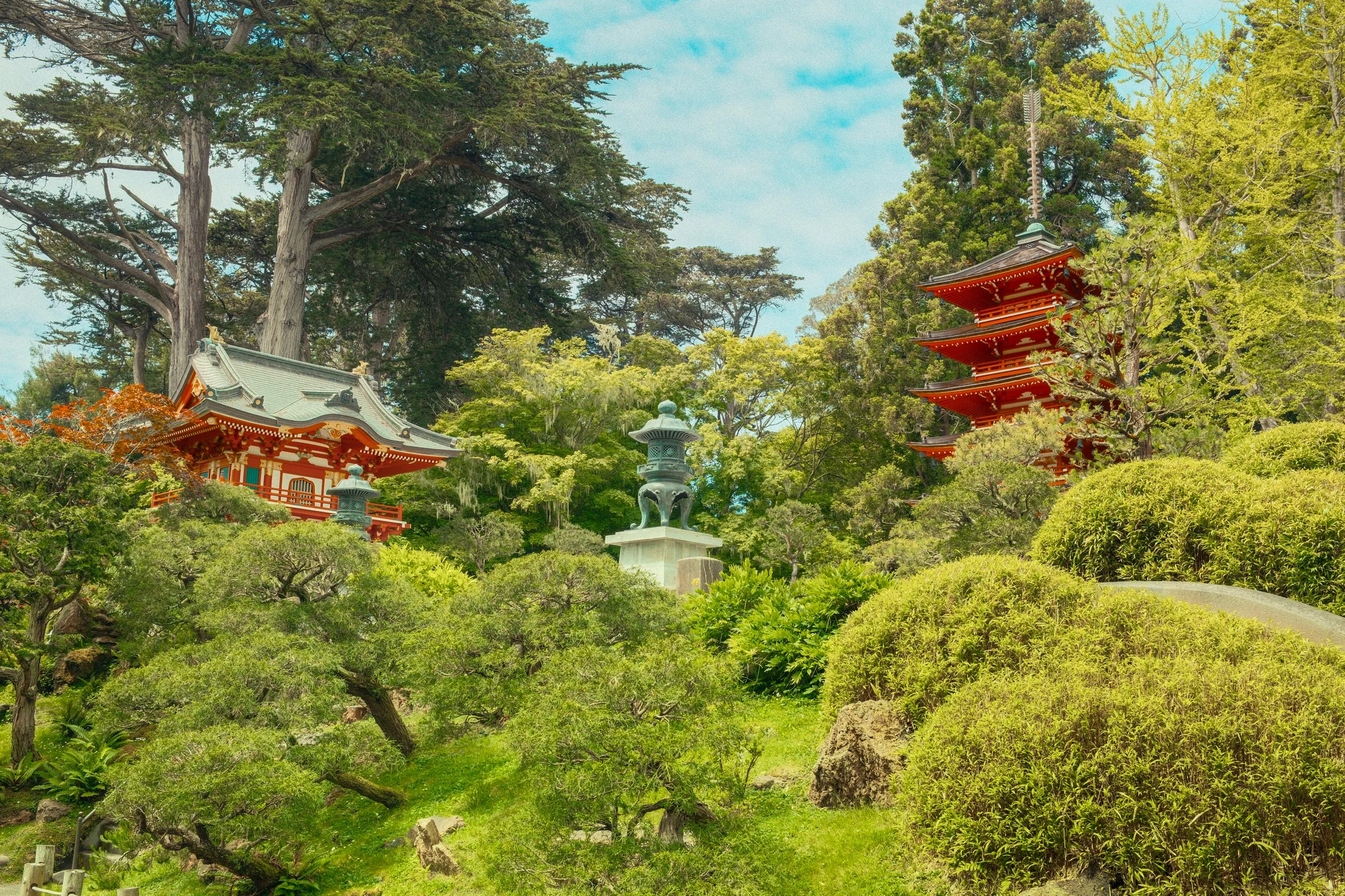 A traditional Japanese garden featuring a red pagoda, a stone lantern, and lush green trees and shrubs.