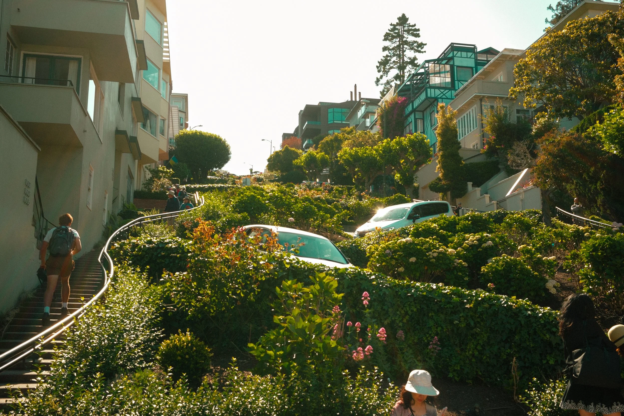 People walking up a steep garden staircase between residential buildings with colorful facades and surrounded by lush flowering plants and trees.