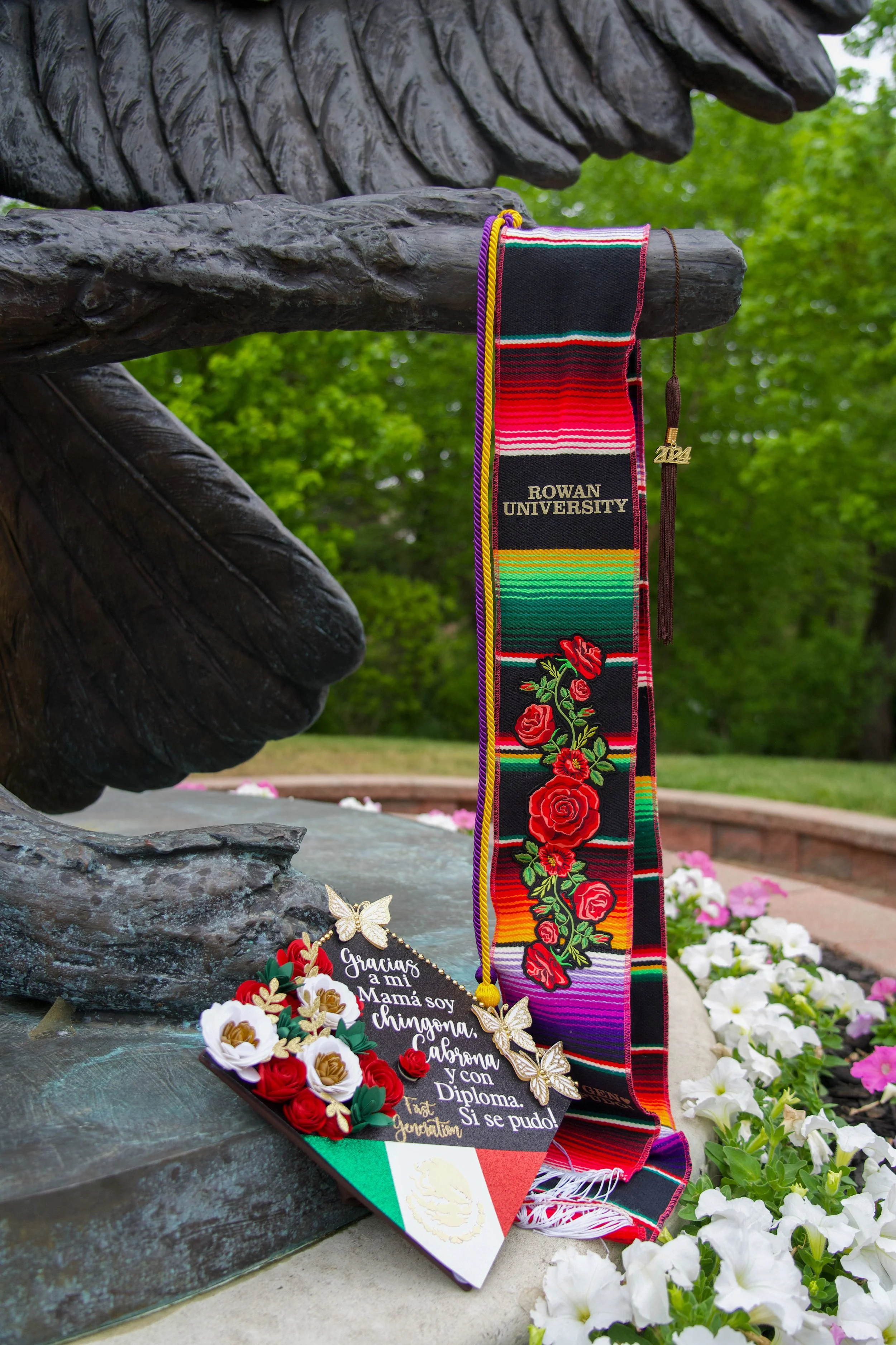 A memorial with a bronze statue featuring a hand and part of a face, adorned with a colorful woven sash from Rowan University and various decorative flowers and a sign in Spanish at the base, surrounded by green trees and flowers.