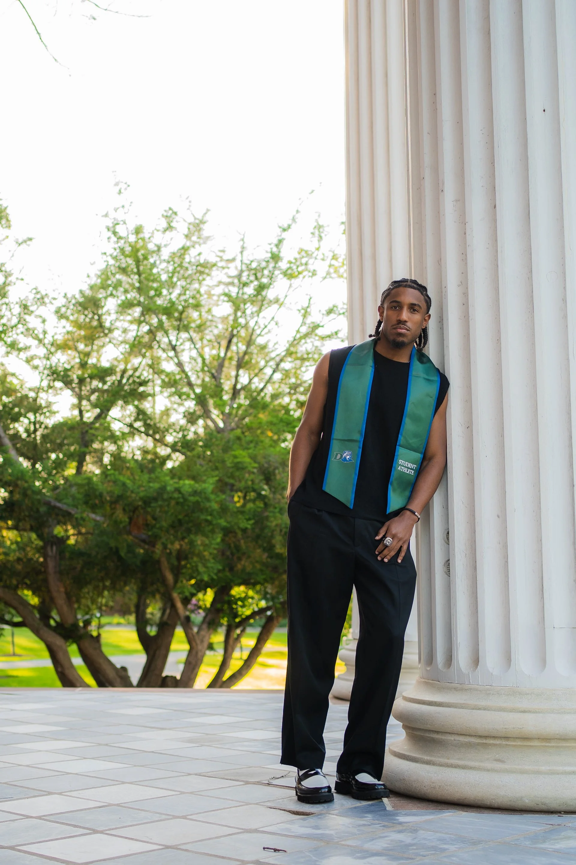 A young man standing outdoors against a large white classical column, wearing black clothing, white and black shoes, and a green and blue student-athlete stole around his neck, with trees and grassy area in the background.