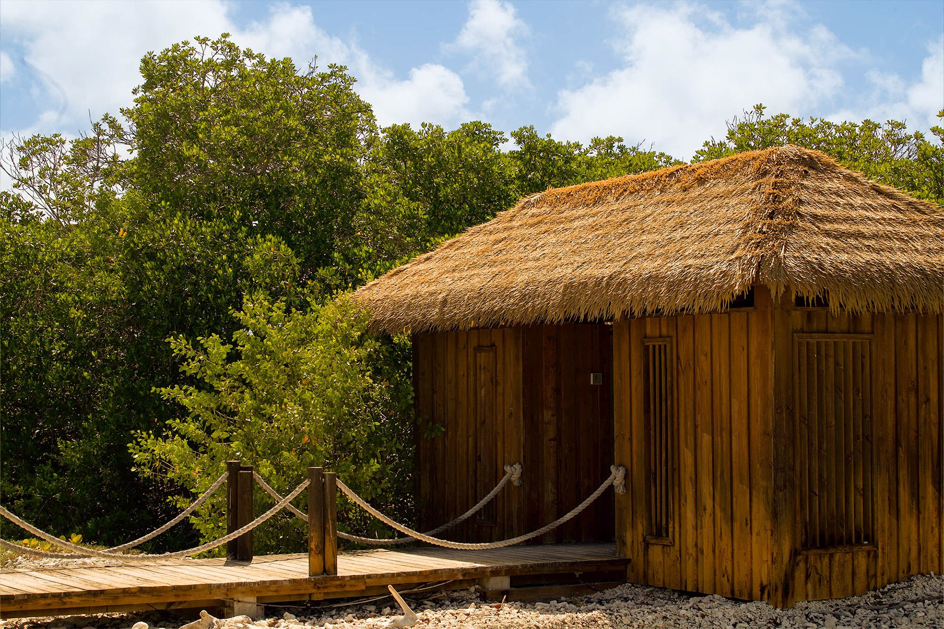 A wooden hut with a thatched roof, surrounded by green trees, under a partly cloudy sky.