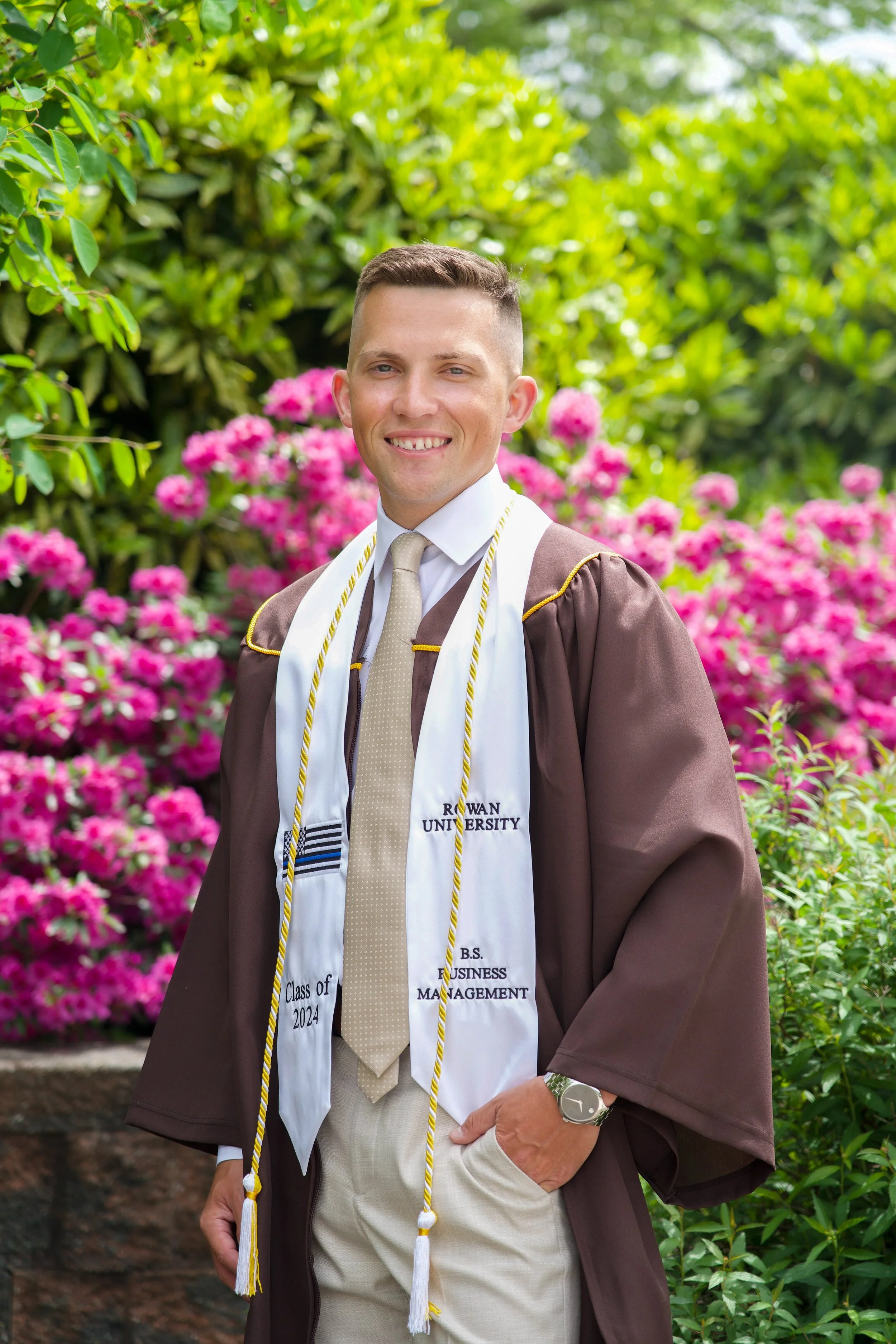 A young man in a graduation gown and cap standing outdoors in front of pink flowers and green foliage, smiling at the camera. He's wearing a cream-colored tie, a watch, and a white stole with university lettering.