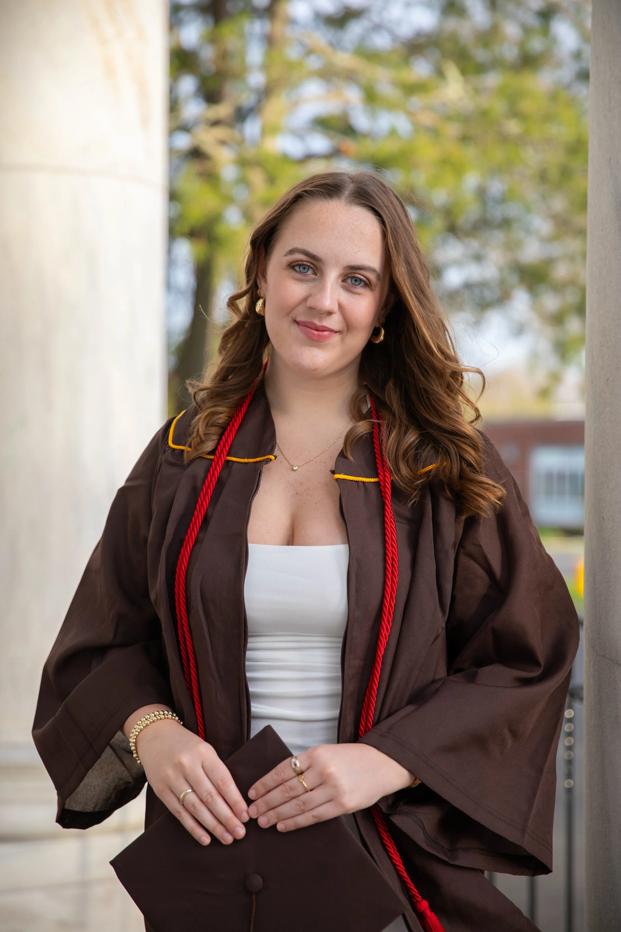 A young woman in a graduation gown and cap held in her hands, standing outdoors between two columns with blurred trees in the background.