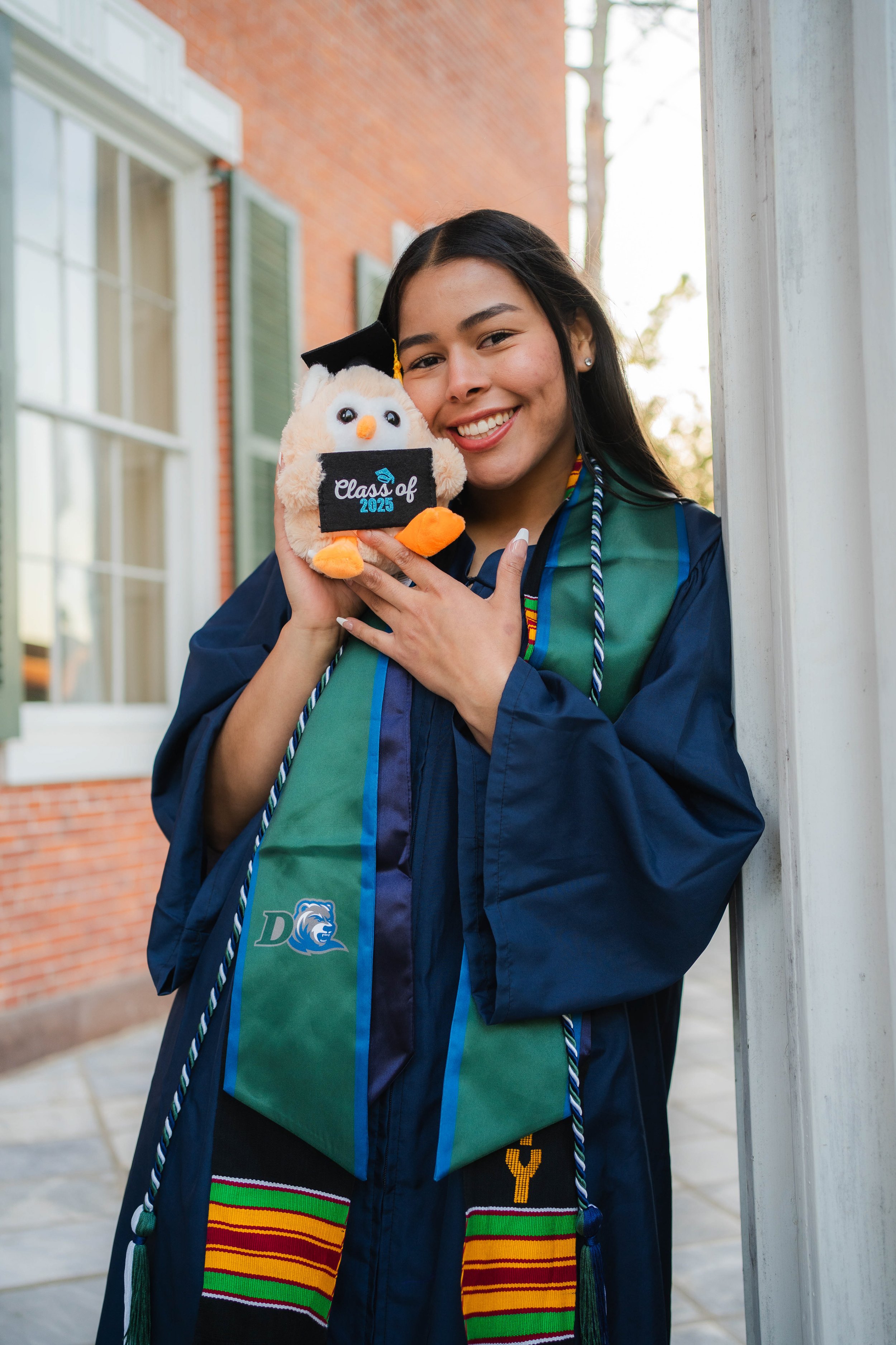 A young woman in a graduation cap and gown holding a plush owl wearing a 'Class of 2025' cap and a Colorado Buffaloes stole, smiling and standing outdoors next to a building with brick walls and a window.