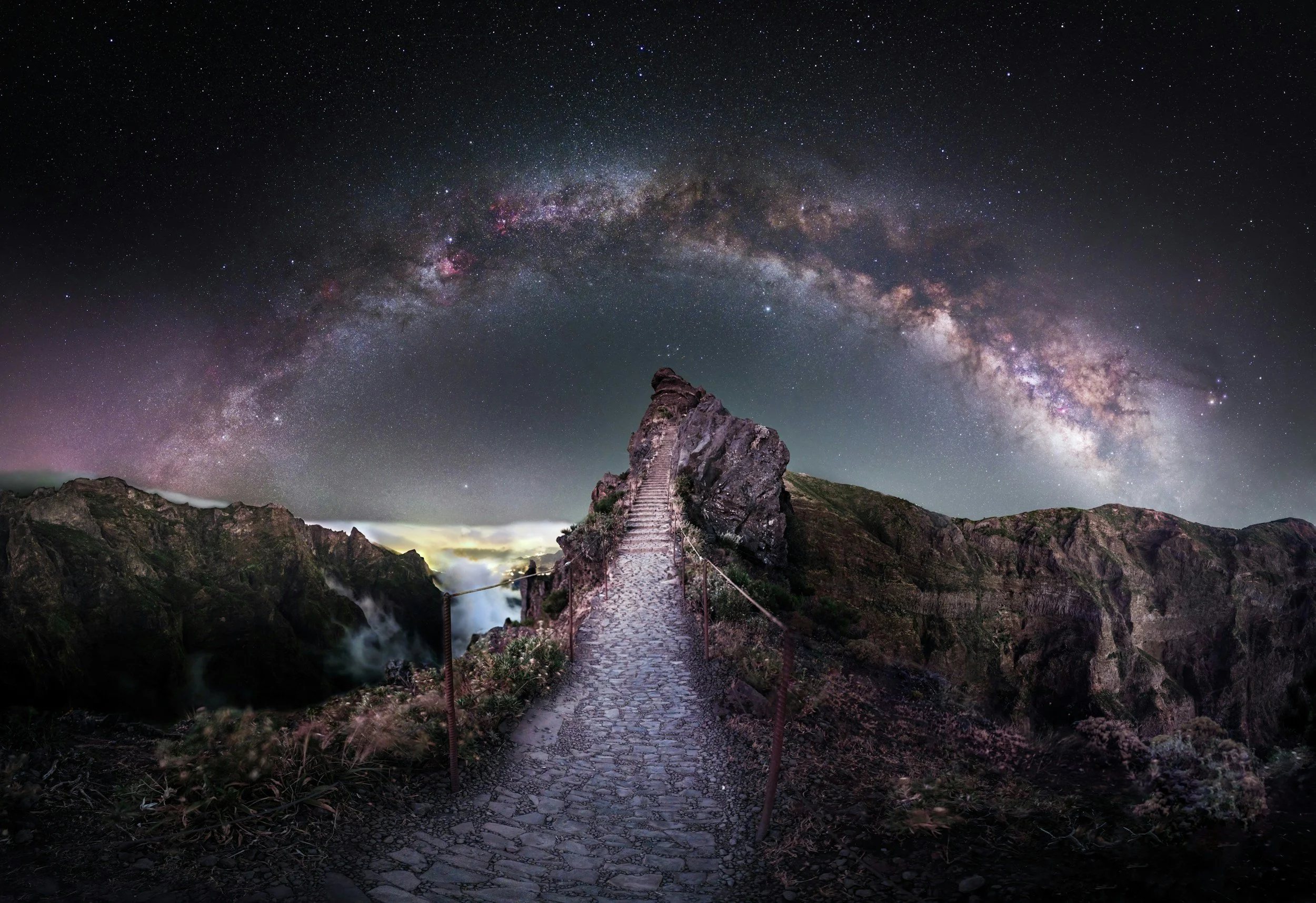 A rocky mountain landscape at night with a stone pathway leading up to a rock formation tower. The sky is filled with stars, and the Milky Way galaxy is prominently visible overhead.