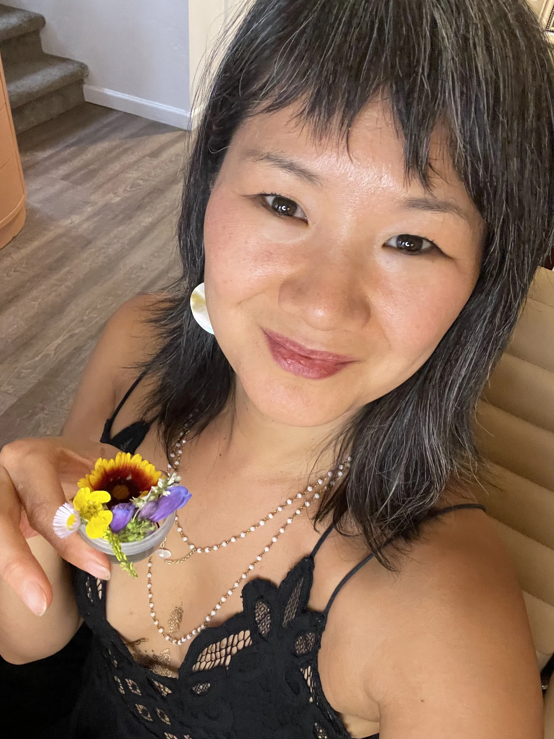 A woman with long dark hair, wearing a black lace dress and pearl necklaces, smiling and holding a small glass with a colorful flower arrangement. She is sitting on a beige cushioned seat with a staircase in the background.
