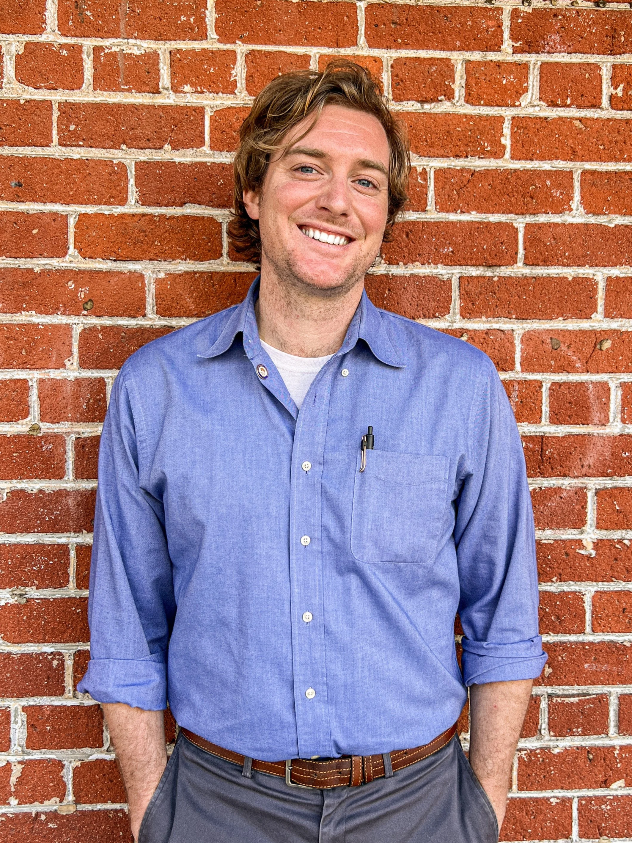 Addison Winslow standing in front of a red brick wall in Chico, California. Addison is smiling and wearing a blue button-up shirt with rolled sleeves.