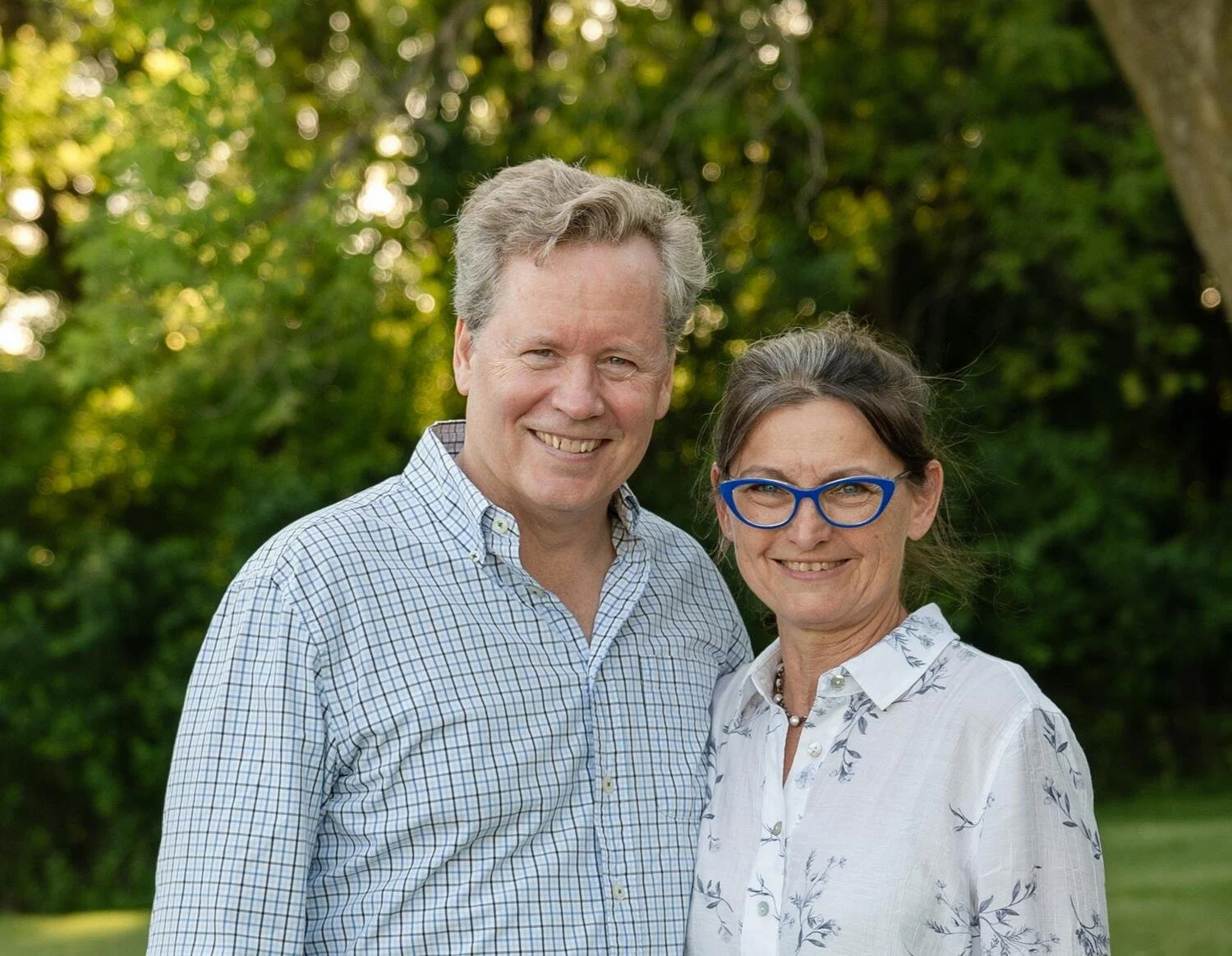 A smiling middle-aged man and woman standing outdoors in front of green trees, both wearing glasses and patterned shirts.