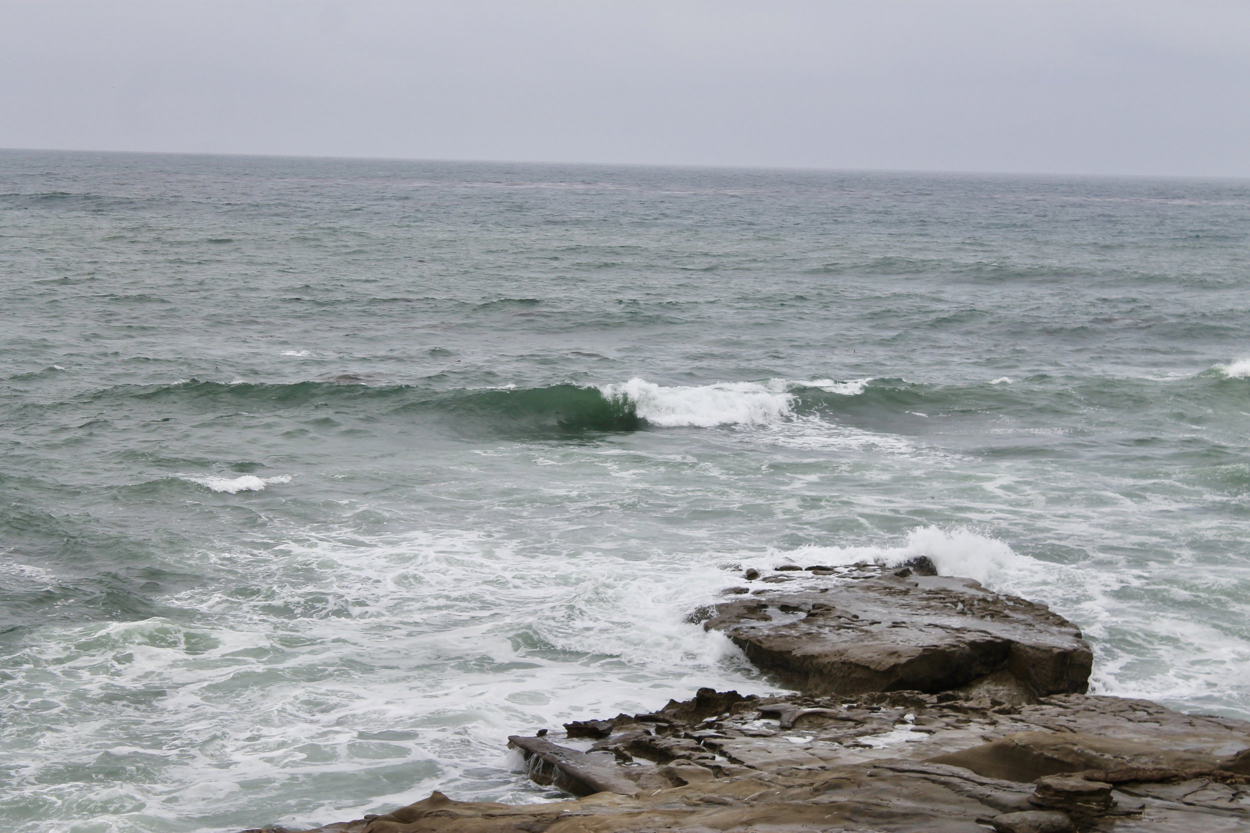 Overcast sky over a calm ocean with small waves, rocks in the foreground.