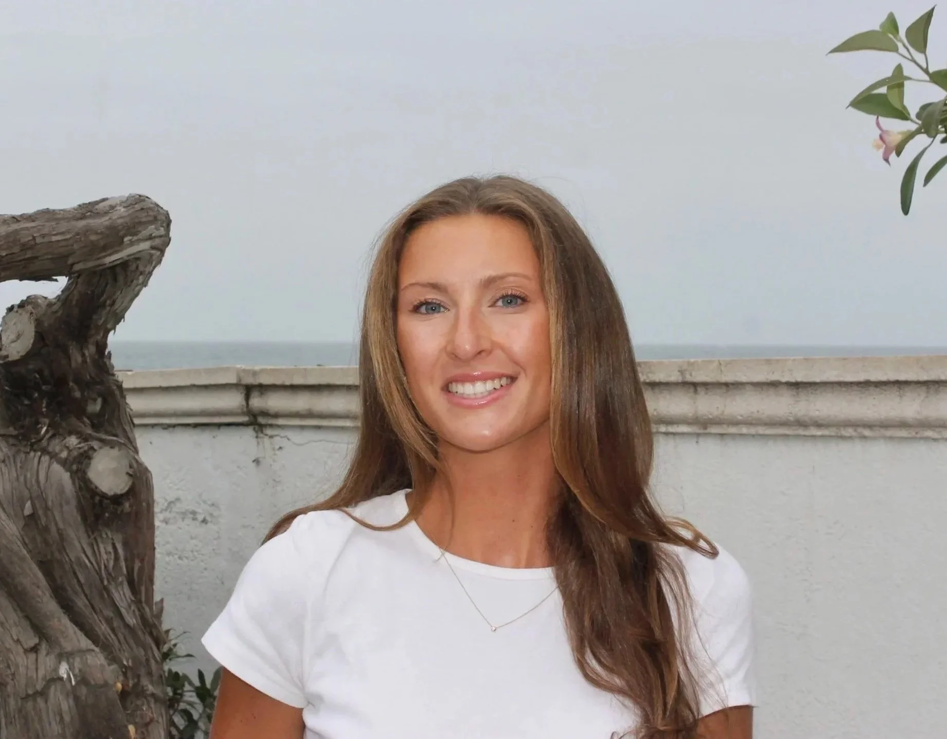 Portrait of a woman with long wavy brown hair, wearing a white shirt and a silver necklace, smiling outdoors near a tree and a white wall, with the ocean in the background.