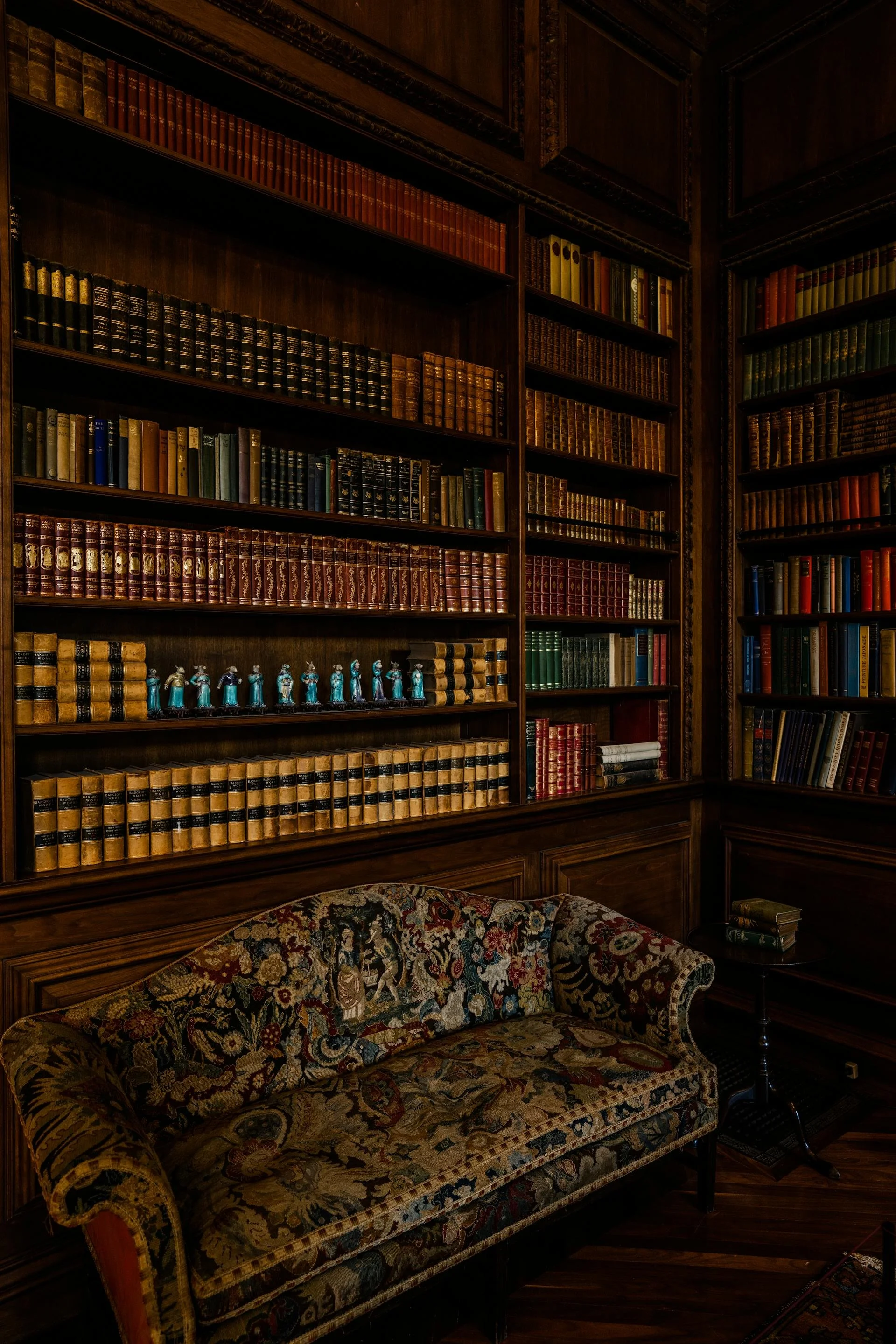 A library with dark wooden bookshelves filled with leather-bound classic books, a floral-patterned upholstered sofa, and a small side table with a lamp in a cozy, vintage setting.