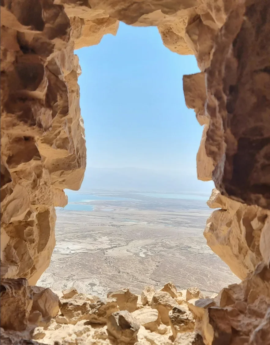 View of a desert landscape with salt flats and a blue sky seen through a rough, irregular opening in a rock wall.