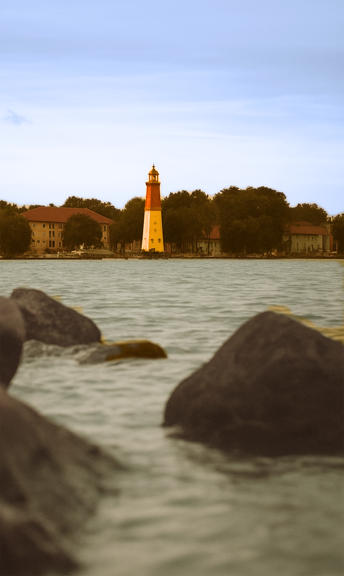 A lighthouse on the opposite side of a body of water, surrounded by trees and buildings, with rocks in the foreground.