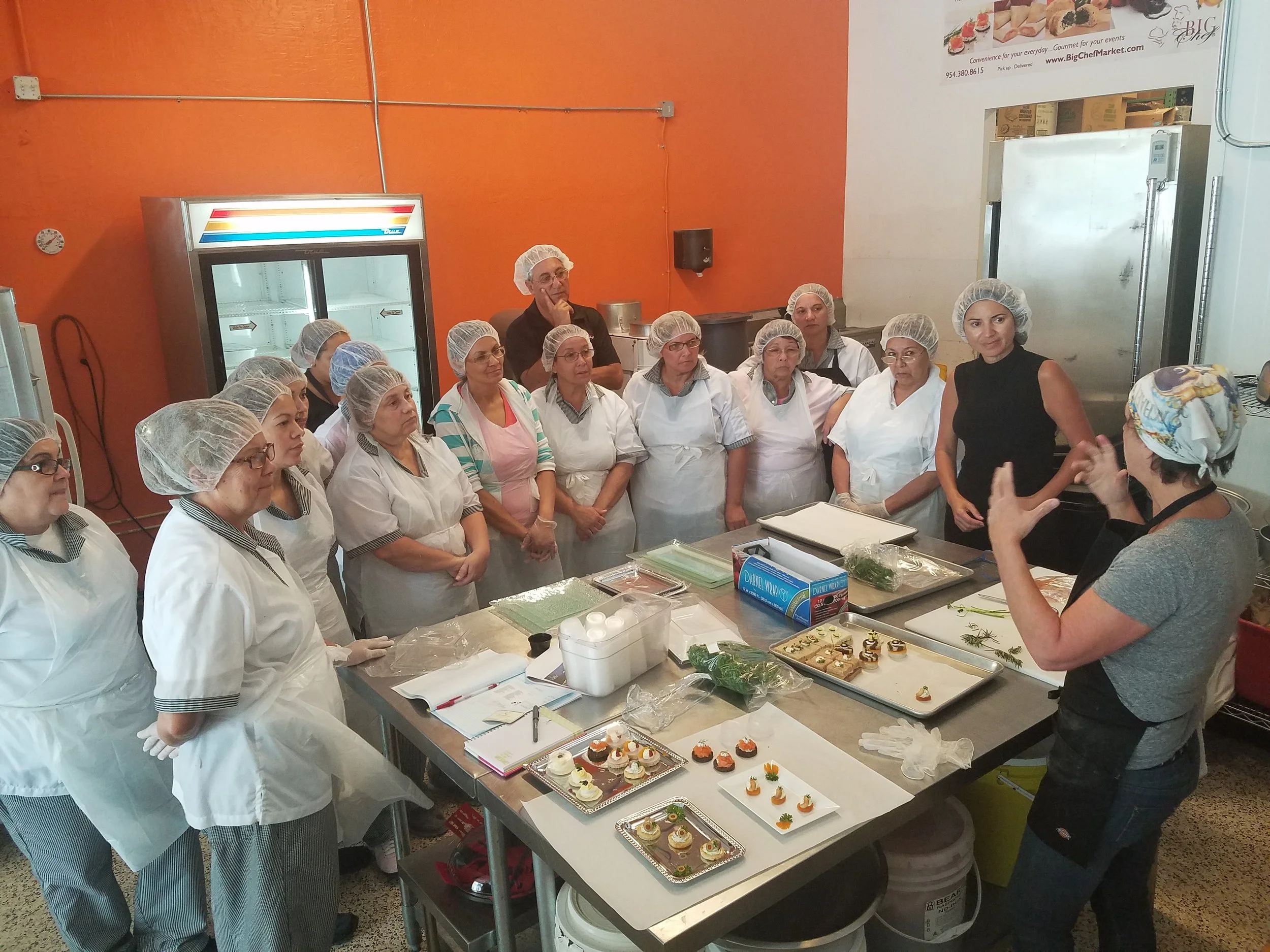 Group of people in hairnets and aprons inspecting decorated cupcakes and pastries on a kitchen table.
