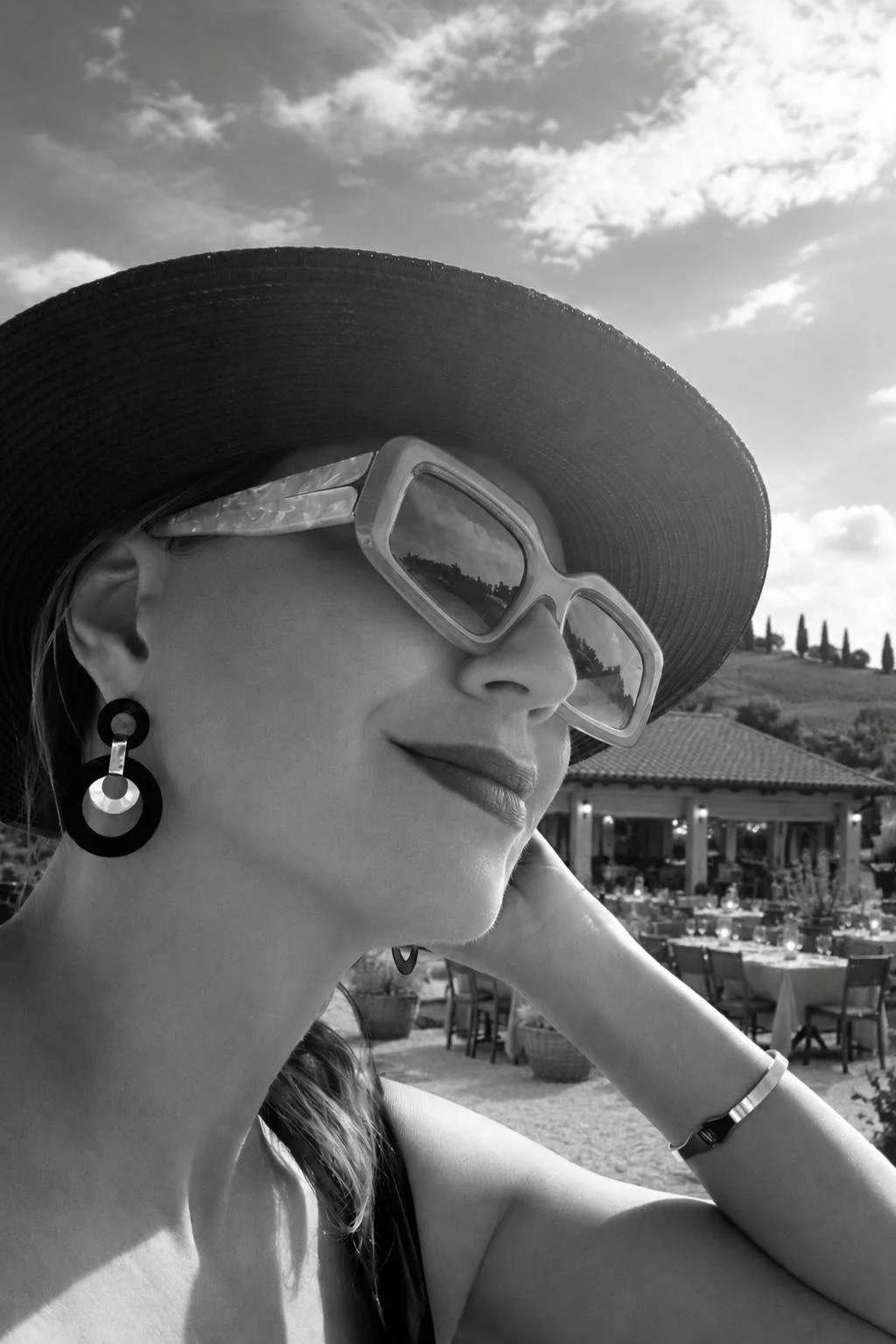 A woman wearing a large sunhat, sunglasses, and earrings enjoying an outdoor setting with tables and chairs, possibly at a restaurant or cafe, with a landscape and cloudy sky in the background, in black and white.